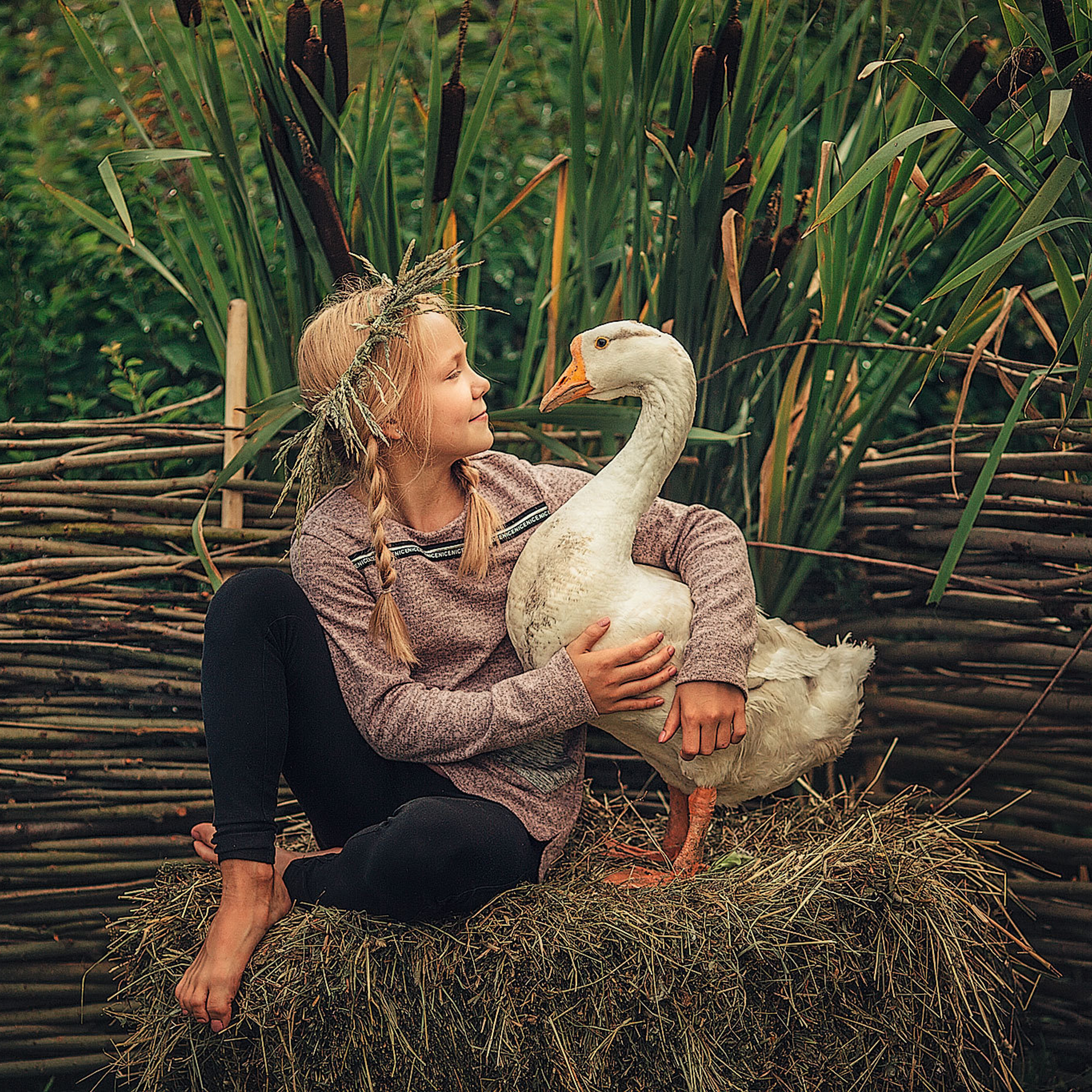 Девочка в венке из цветов на голове обнимает гуся и смотрит на него