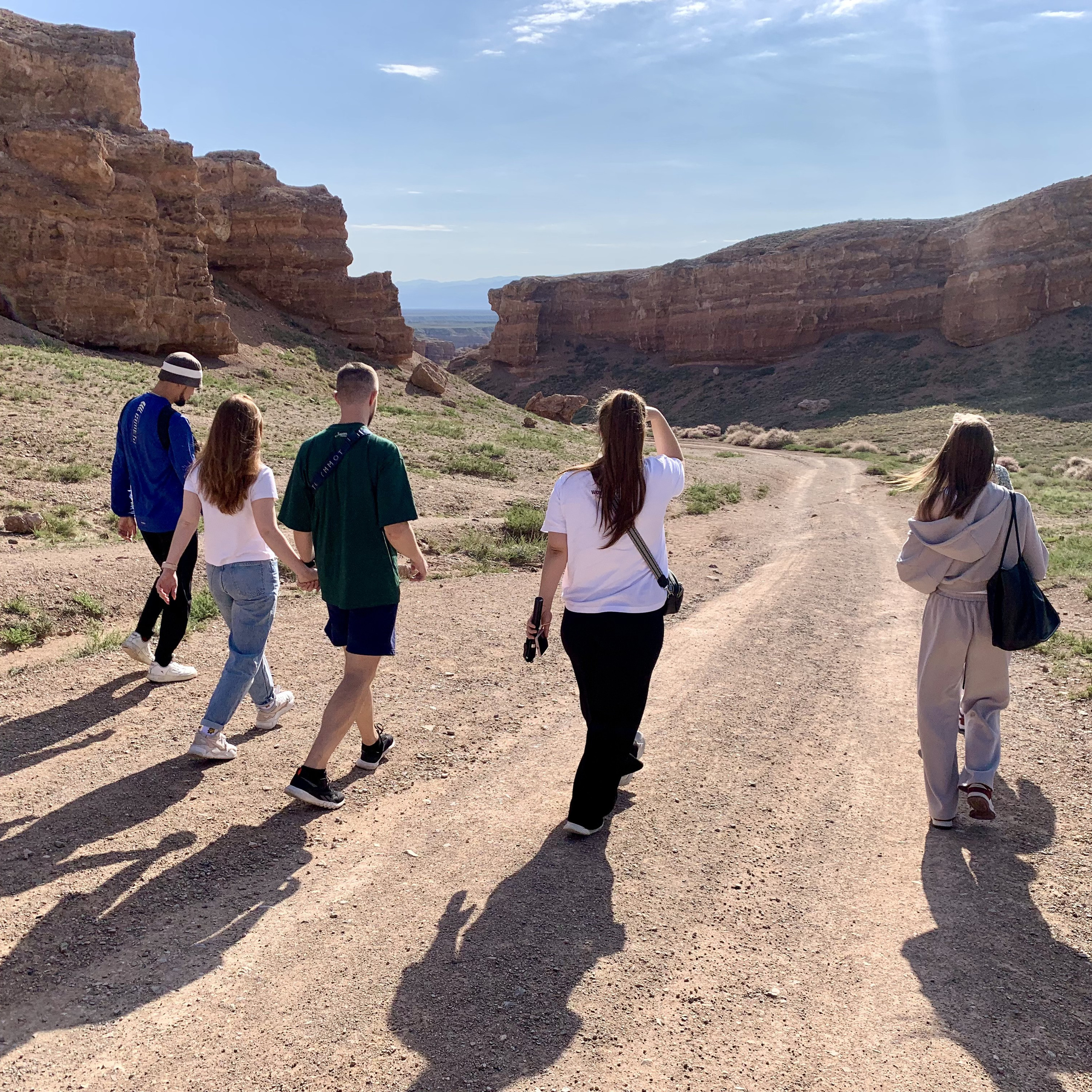 Группа людей идёт по грунтовой дороге в направлении Долины Замков. На заднем плане видны высокие скалы Чарынского каньона с оранжево-коричневым оттенком, окружающие тропу. Небо ясное с небольшими облаками, а солнечный свет создаёт тени от людей. Вокруг расстилается пустынный ландшафт с редкой зеленью.