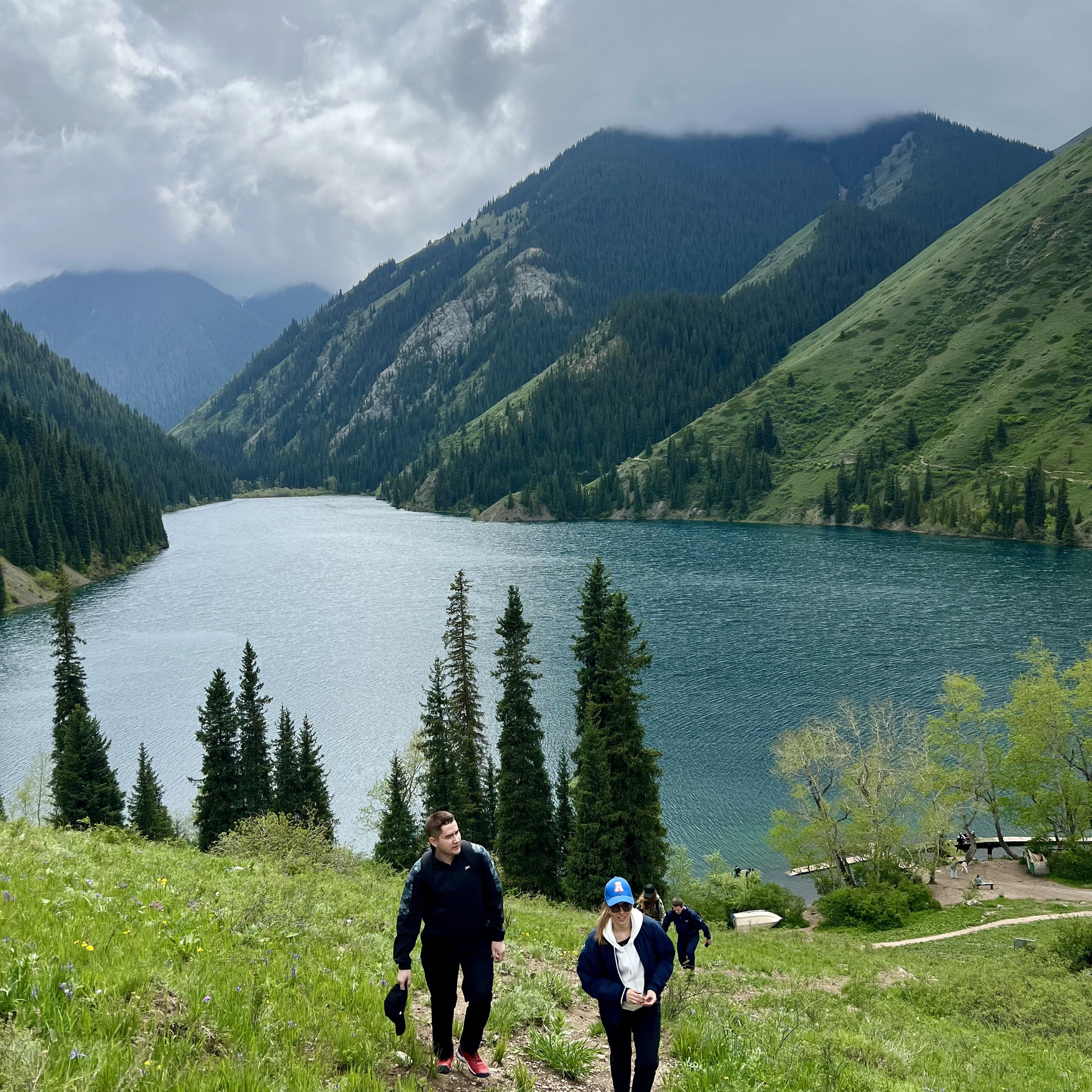 Люди поднимаются по склону холма с видом на красивое горное озеро Кольсай. Озеро окружено высокими зелеными холмами и еловыми деревьями. Над горами нависают облака, создавая атмосферу умиротворения и свежести.