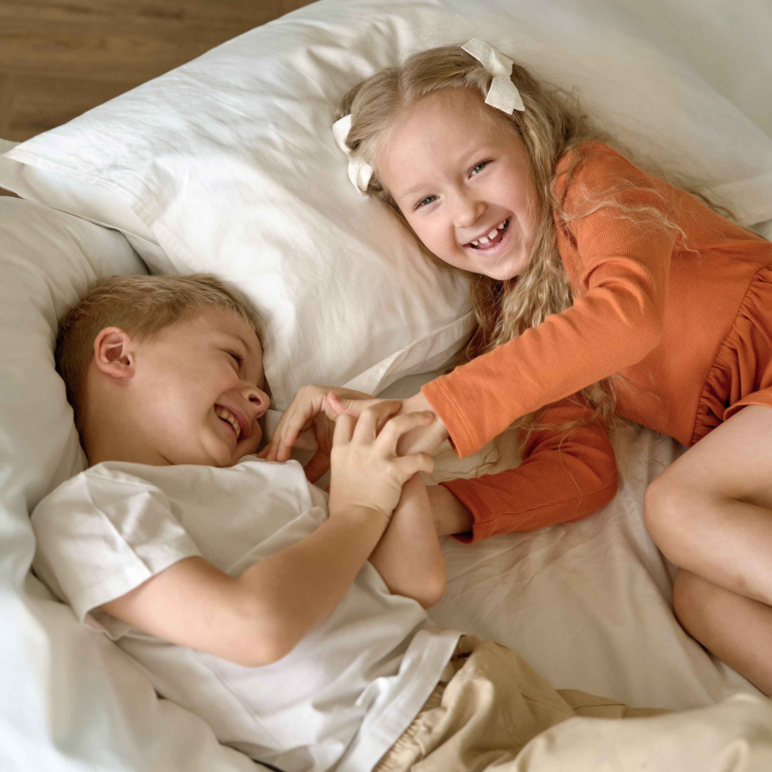 Счастливые дети играют на кровати: брат и сестра смеются, держась за руки. Авторская семейная фотосессия в уютной спальне