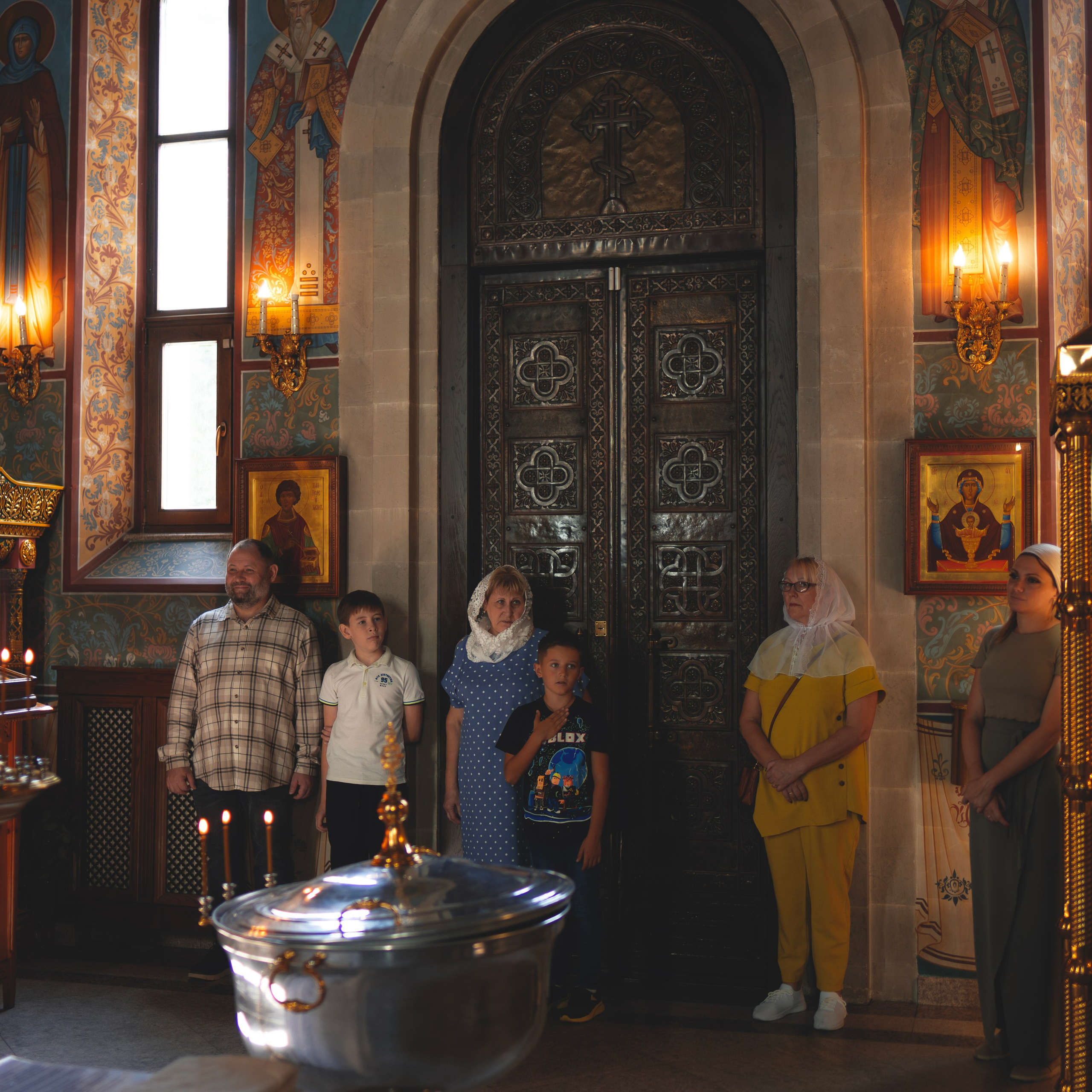 Группа людей стоит в православной церкви перед массивными резными дверями. Среди них взрослые и дети, одетые в светлую и скромную одежду. На некоторых женщинах платки. Интерьер церкви украшен иконами, деревянными резными элементами и свечами, создающими теплое освещение. На переднем плане видна серебристая купель, подготовленная для обряда крещения. Атмосфера в церкви спокойная и торжественная.
