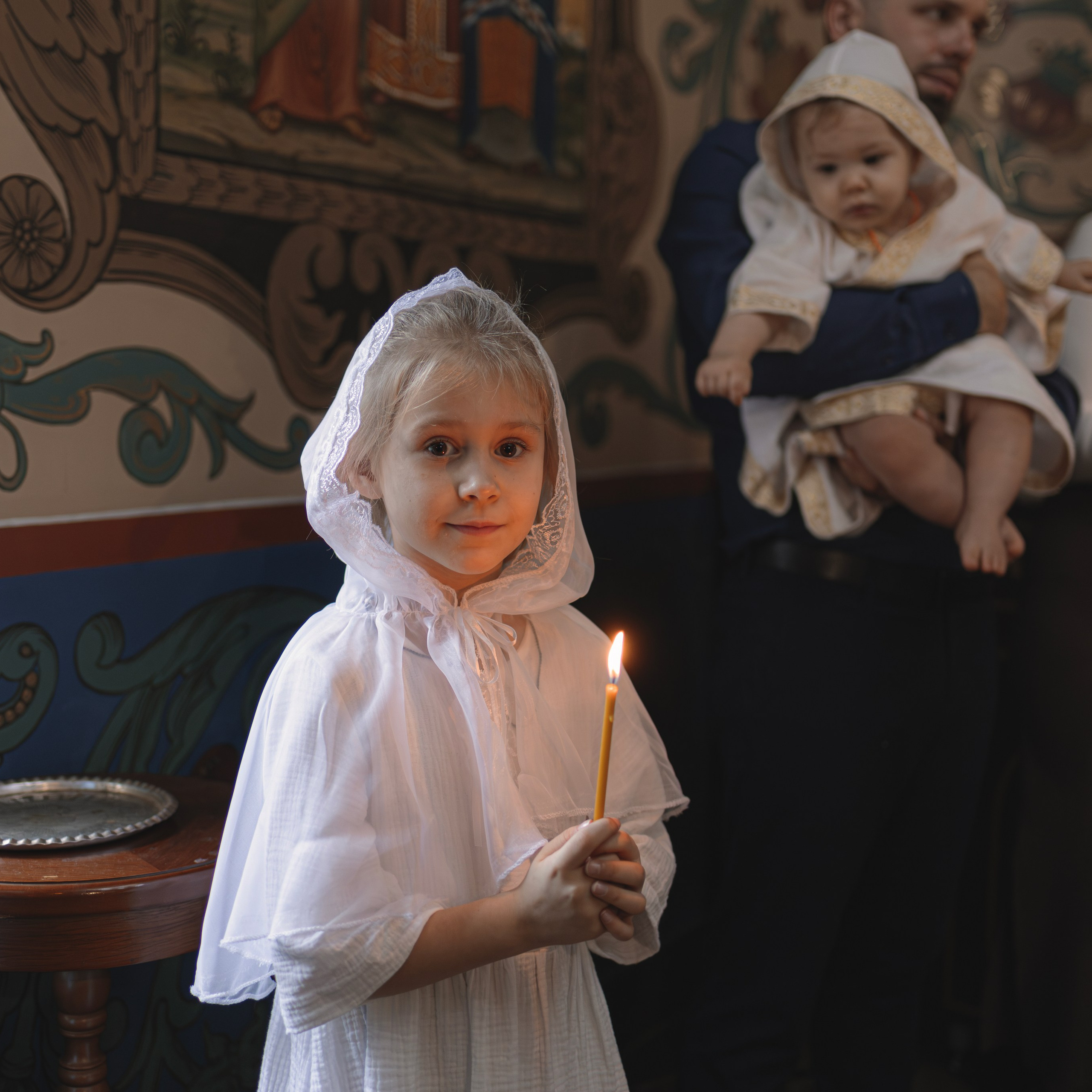 Девочка в белом головном уборе с зажжённой свечой в руках символизирует чистоту и свет веры. На заднем плане видны члены семьи, которые с благоговением держат младенца, что добавляет кадру чувство близости и единства. Нежное освещение и традиционные росписи на стенах храма создают ощущение уюта и торжественности момента.