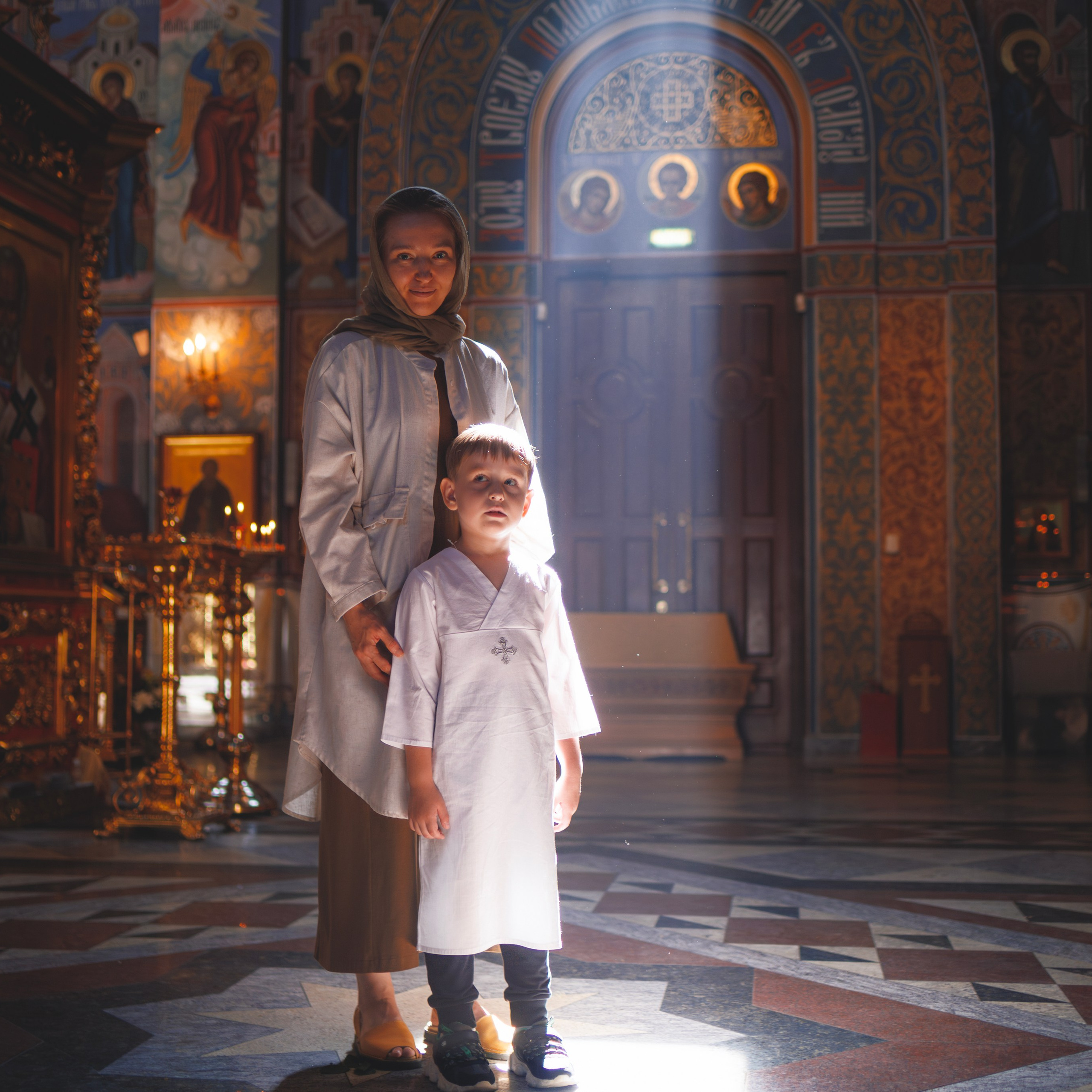 Женщина и мальчик стоят в православной церкви, освещённые лучом света, падающим сверху. Мальчик одет в белую крещальную рубашку, а женщина — в светлую одежду и платок. Вокруг них виден богатый интерьер храма с золотыми украшениями, иконами и сложными орнаментами на стенах. Атмосфера в церкви торжественная и спокойная, подчёркивающая значимость момента.
