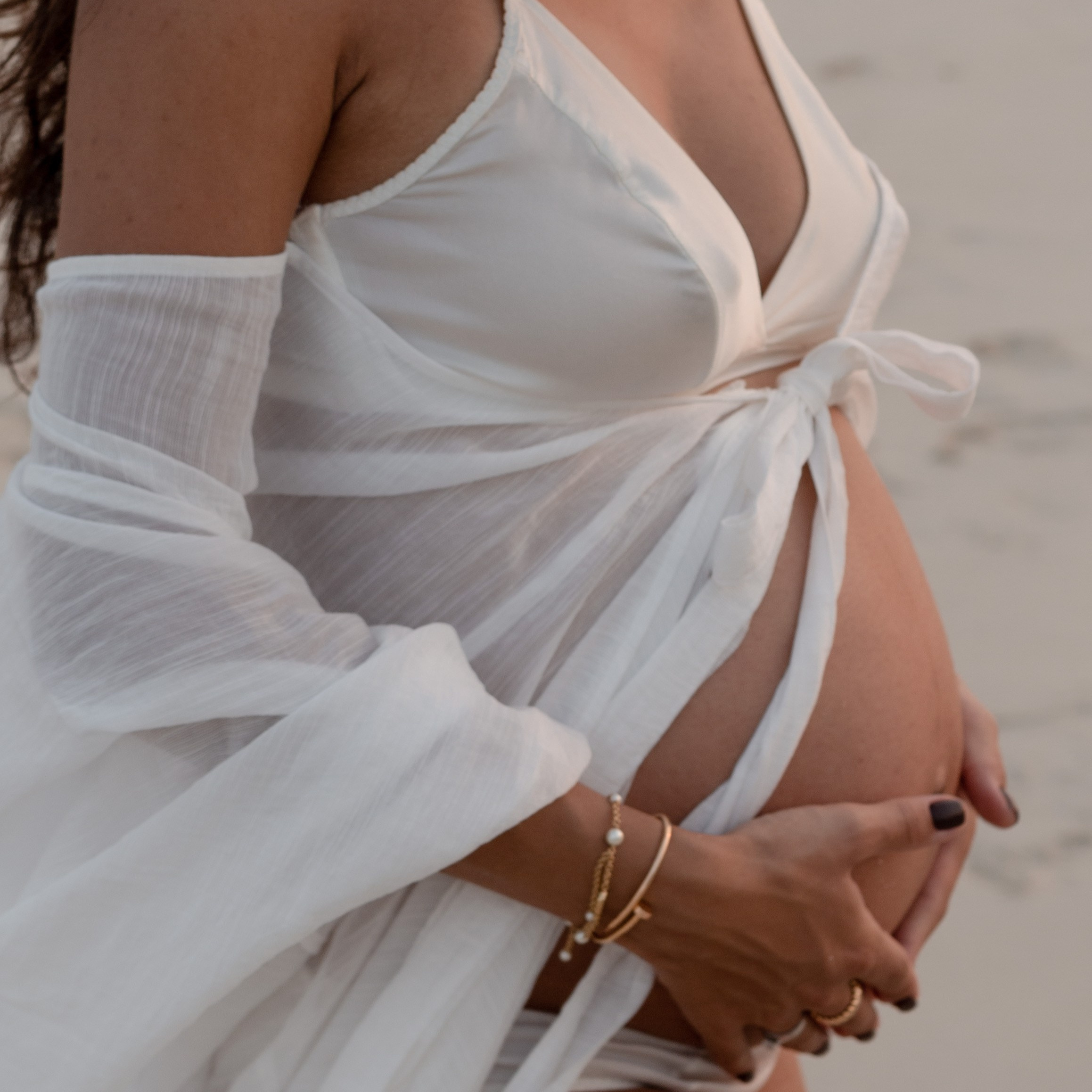 A close-up of a pregnant woman in a white outfit standing on the beach, gently holding her belly and wearing delicate jewelry