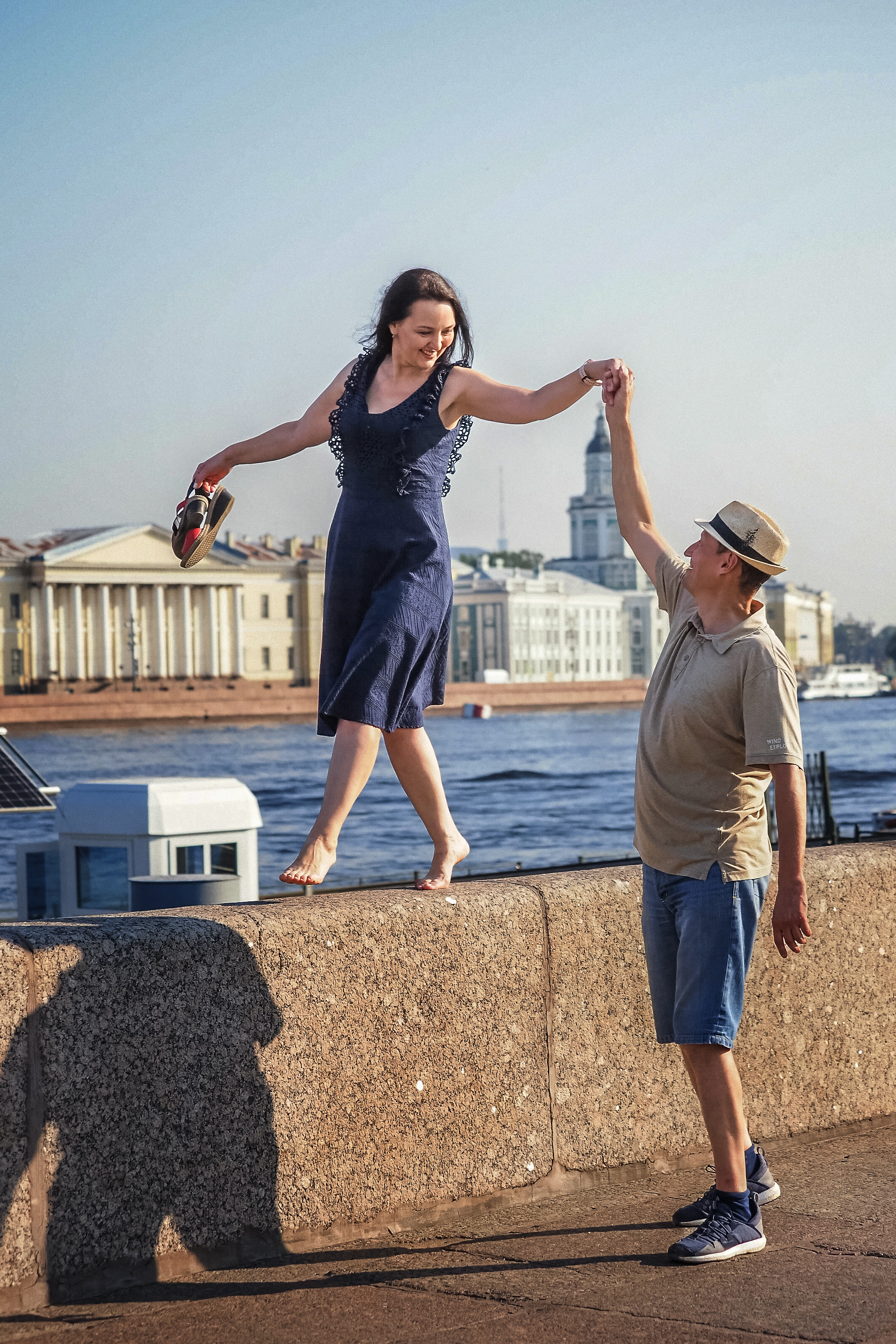 мужчина держит за руку женщину, которая идет босиком по каменному бортику набережной реки Невы в Санкт-Петербурге