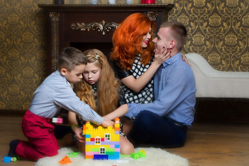 молодая семья. рыжеволосая мама обнимает папу. дети: мальчик и девочка играют в конструктор