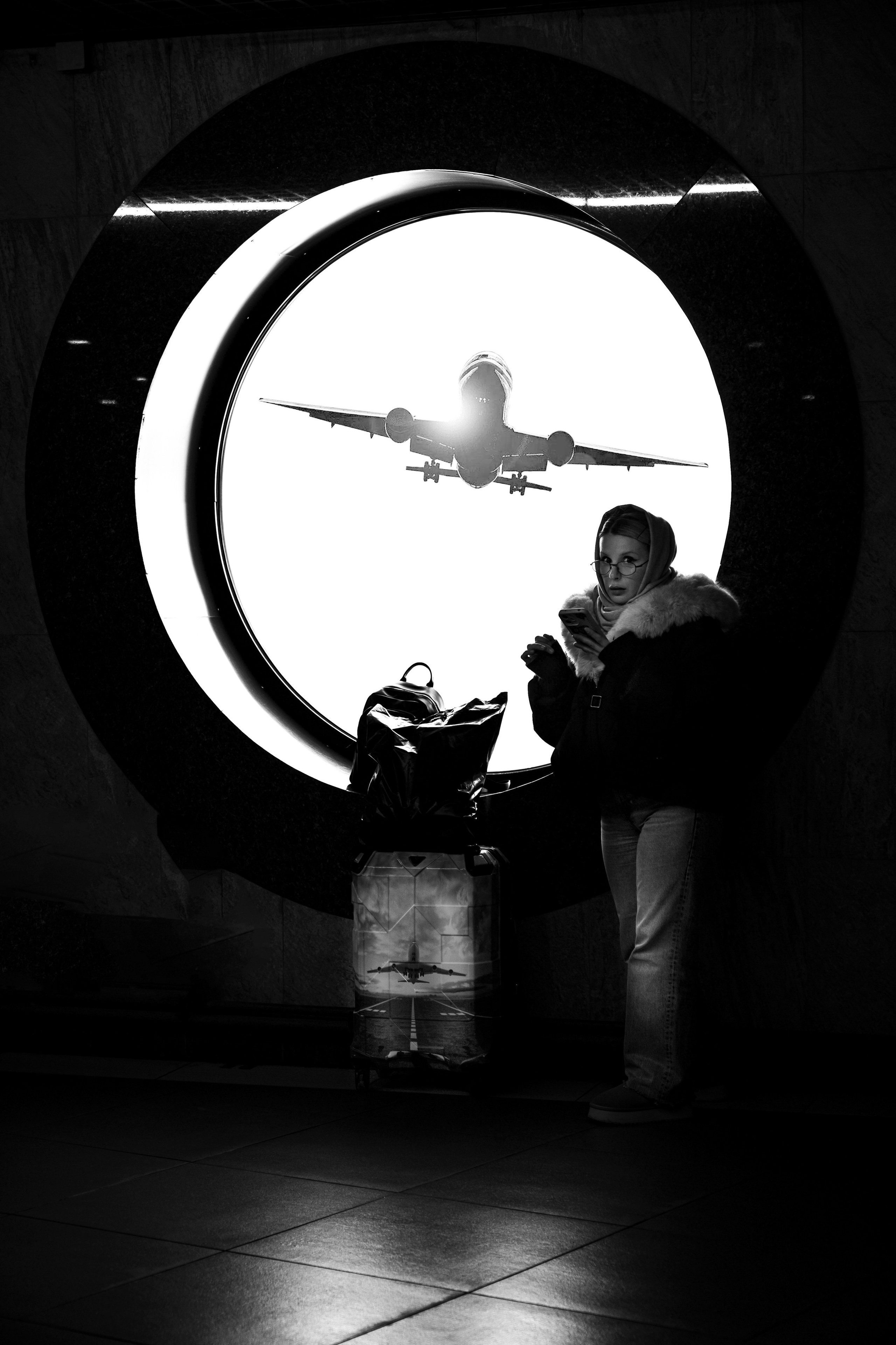 Девушка в платке с телефоном на фоне огромного круглого окна и взлетающего самолёта. Чёрно-белая стрит-фотография Мальцева Евгения.