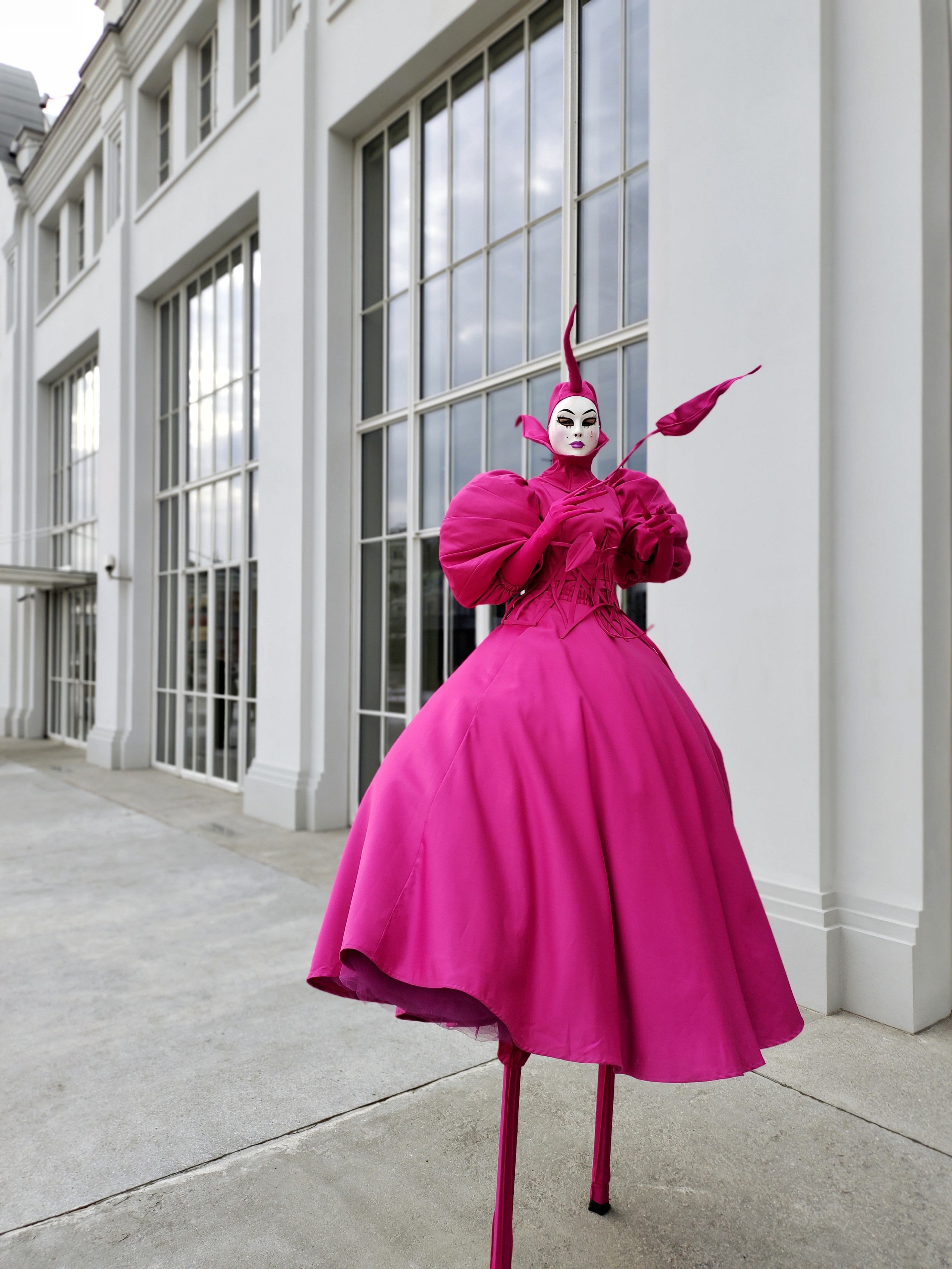 Ходулист к костюме цвета Фуксия встречает гостей у белого здания.