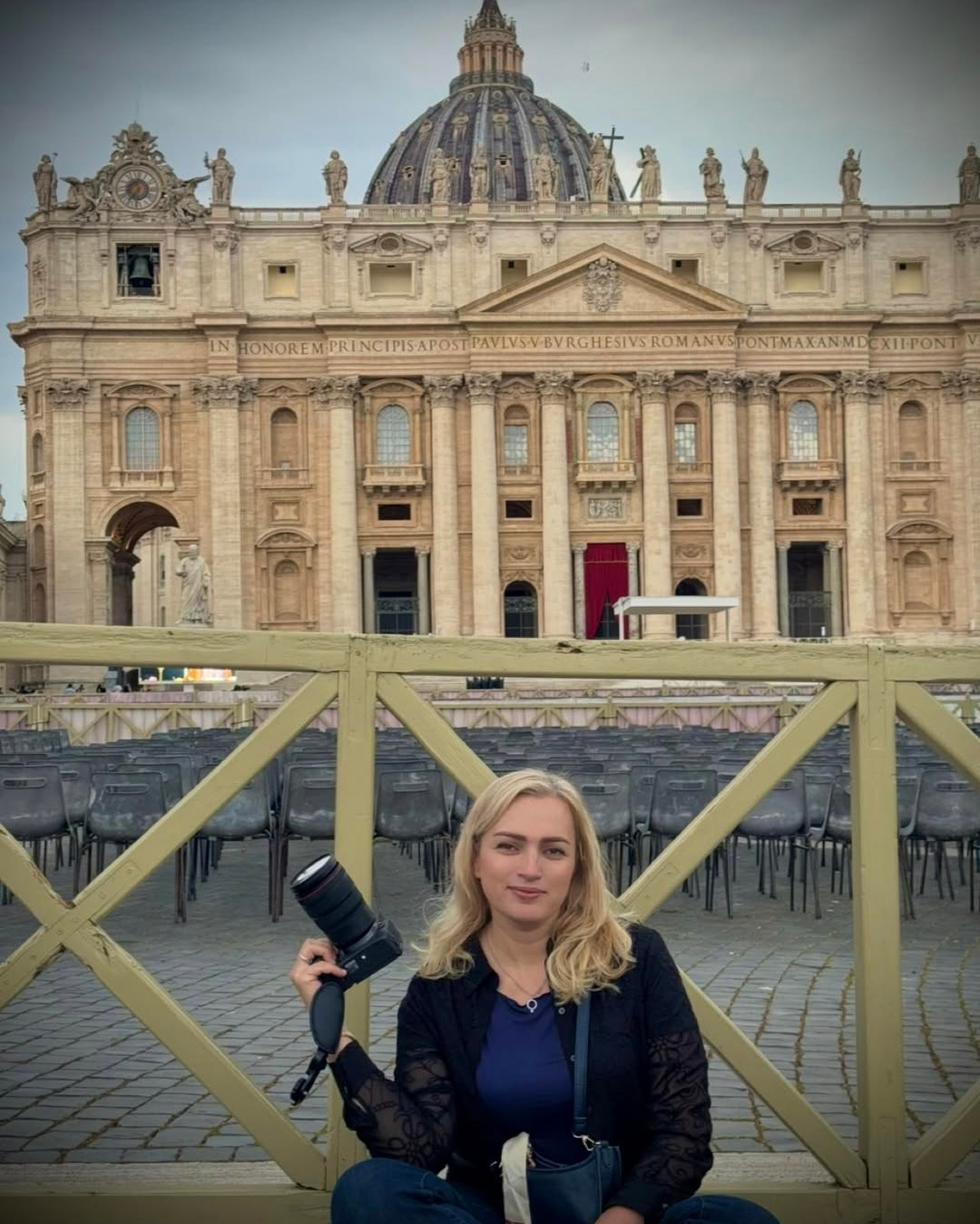 Юлия Перфетти-фотограф сидит с камерой на площади Святого Петра, за ней — величественный фасад и купол собора Святого Петра в Ватикане.