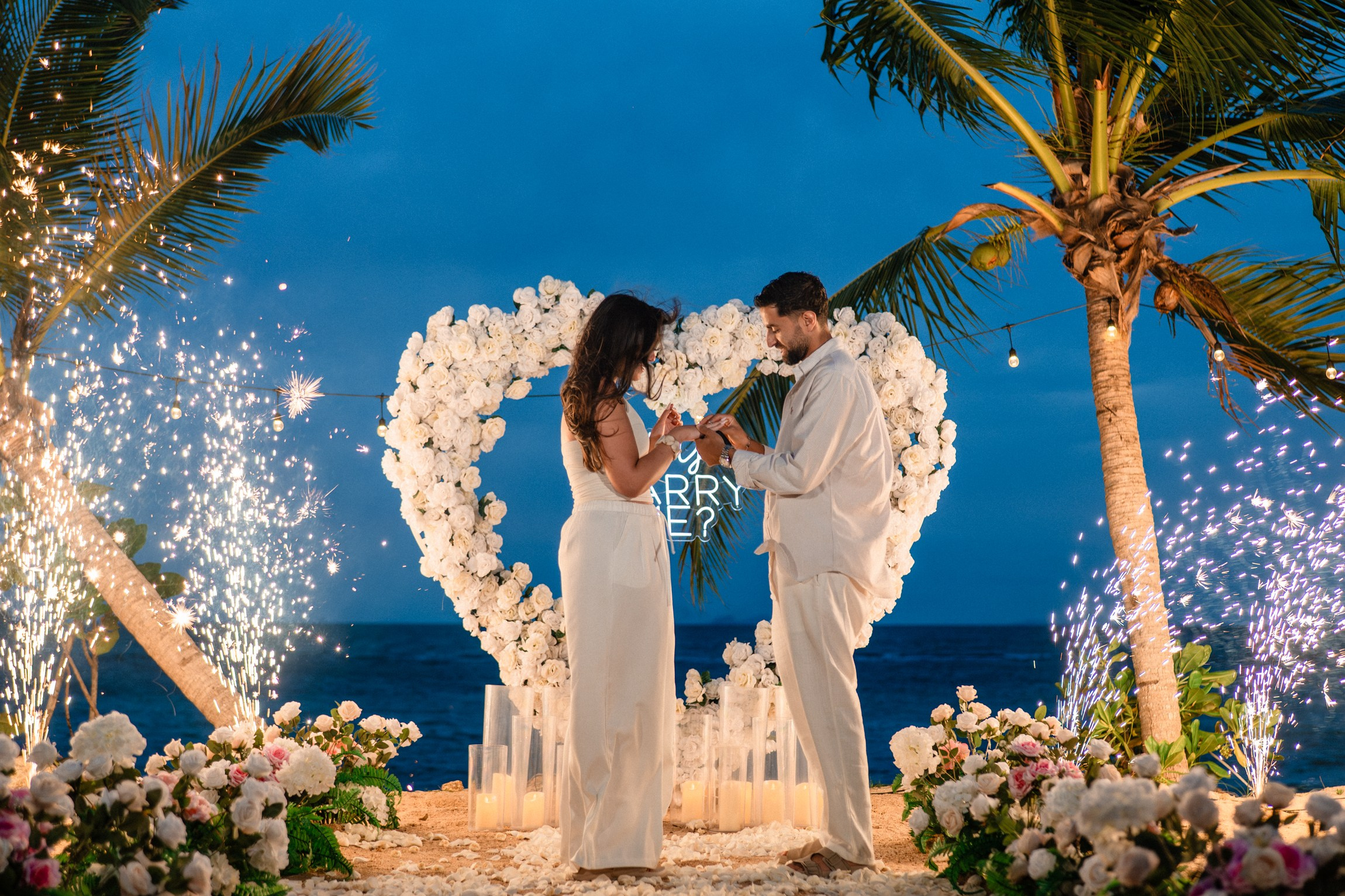 Romantic beach proposal in Koh Samui with a couple under palm trees, white flowers and sparkling fireworks at night.