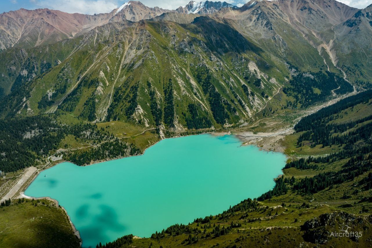 Вид на Лунный Каньон реки Чарын недалеко от Алматы, окружённый зелёными деревьями и кустарниками, на фоне красочных скал в солнечный день. На переднем плане женщина сидит на утёсе и любуется пейзажем