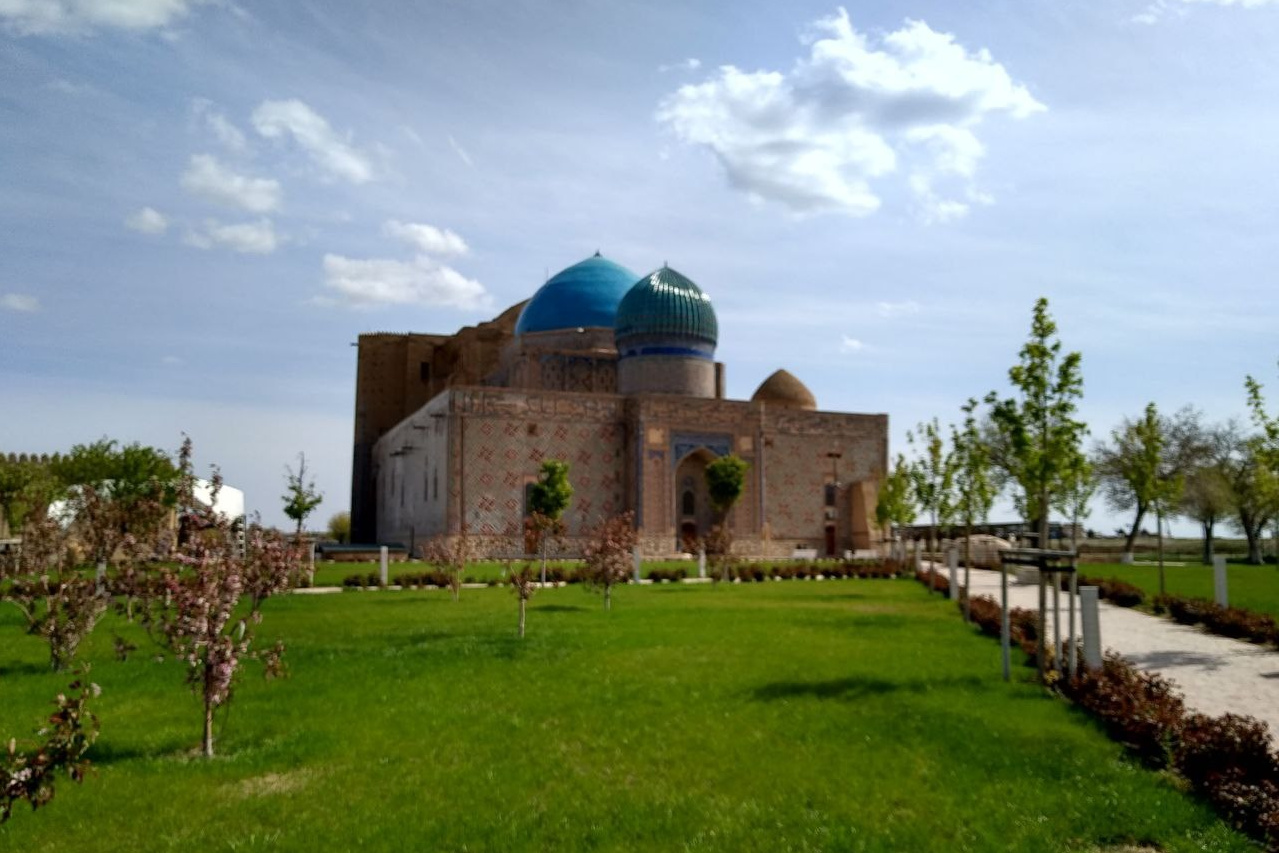 Историческое здание мавзолея Ходжи Ахмеда Ясави в Туркестане, Казахстан. Стены украшены орнаментами, а купола ярко голубого цвета выделяются на фоне неба с лёгкими облаками. Зелёный ухоженный газон и деревья окружают строение, создавая спокойную и гармоничную атмосферу. Дорожка ведёт к зданию, добавляя перспективу в кадре.