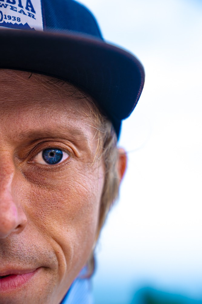 Мужчина в белой рубашке и джинсовых шортах стоит на фоне зелёных деревьев и жёлтого кустарника. Он смотрит прямо в камеру, руки в карманах, поза расслабленная. Сцена выглядит спокойной и яркой благодаря дневному свету и насыщенным цветам природы.