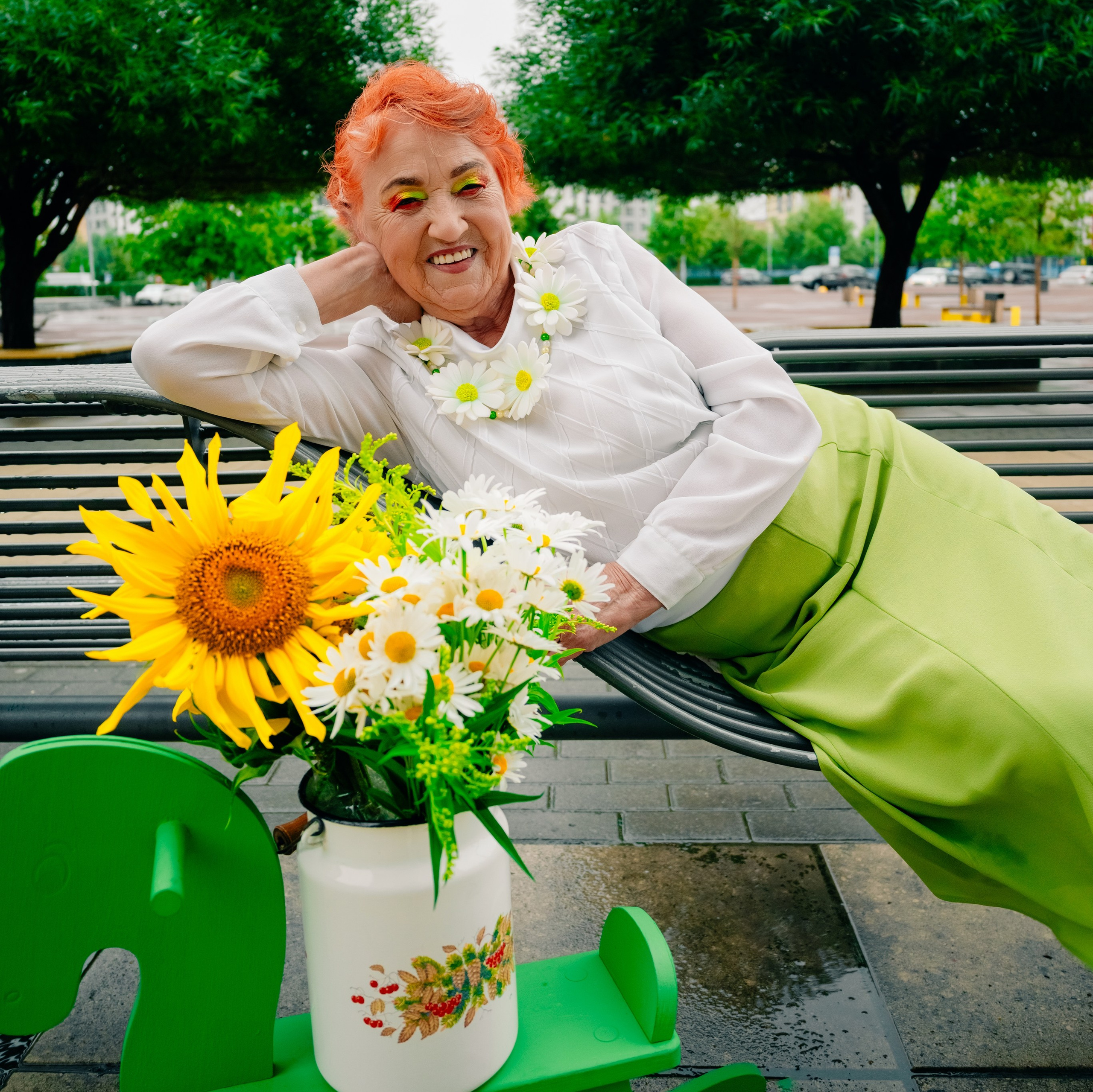 бабушка сидит на лавочке рядом с деревянной лошадкой и букетом ромашек в бидоне