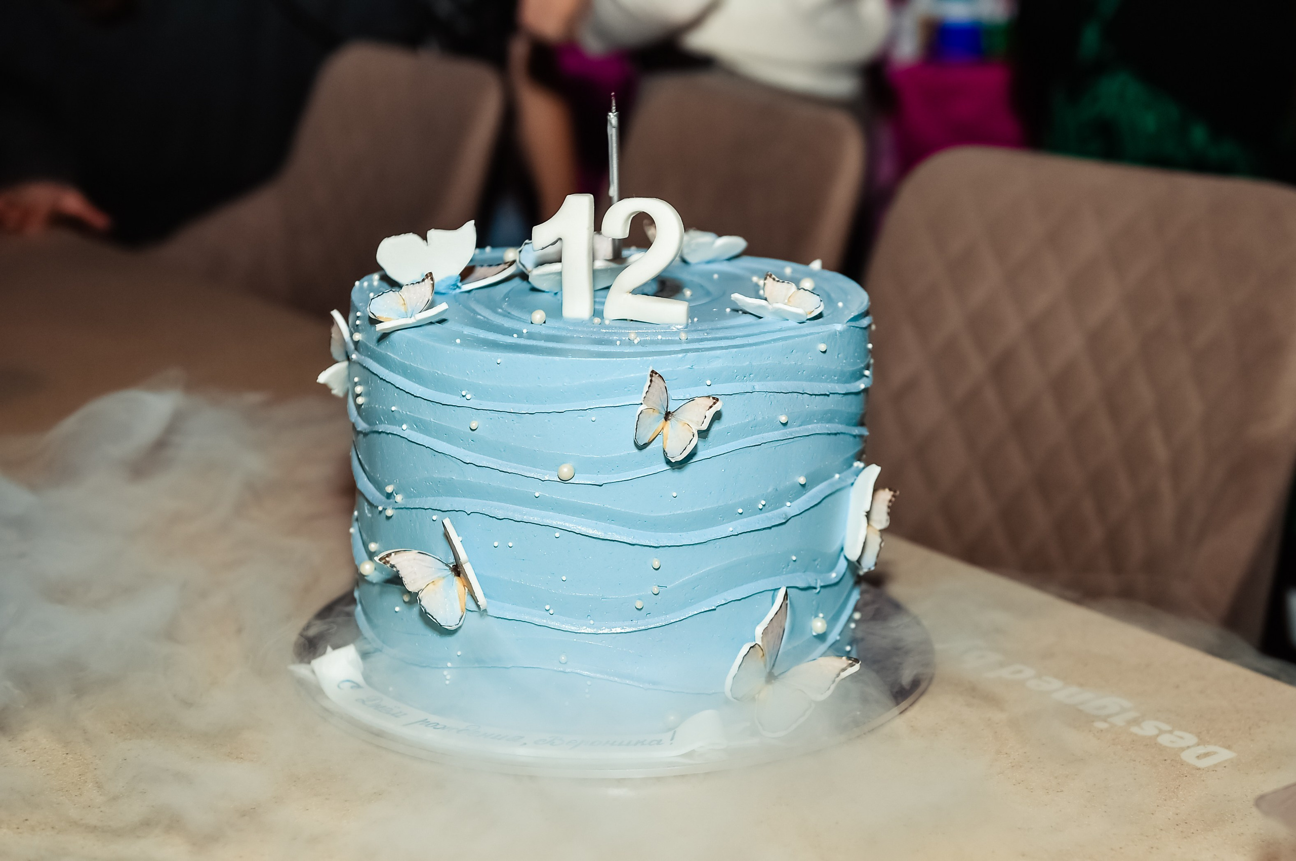 Репортажная фотография, на которой изображен красивый торт с голубой мастикой, декорированный белыми бабочками. На торте стоит одна свеча и цифры 12