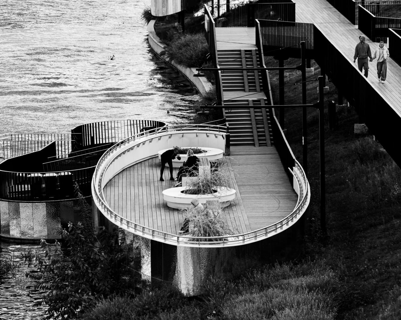 Черно-белая фотография изогнутой набережной с деревянным мостом и людьми в разных позах: двое общаются, один сидит, пара идет по мосту. Река слева, растительность и туман создают атмосферу таинственности и глубины. Городская фотография Вадима Топорёнка, отражающая концепцию археологии душ и диалога с городом в проекте "Манифест Теней". Изогнутая дорожка символизирует путь поиска, а люди — живые истории, которые город хранит в своих трещинах. Контраст черно-белой палитры подчеркивает глубину эмоций и связь между прошлым и настоящим. Фотография, которая не просто фиксирует момент, а ловит то, что остается после нас — в тенях, в ритме городского дыхания и в диалоге между человеком и пространством. Снимок, воплощающий философию фотографа, где каждый кадр — это свидетельство того, что город не фон, а живой собеседник, который говорит через трещины в асфальте и шепчет через туман.