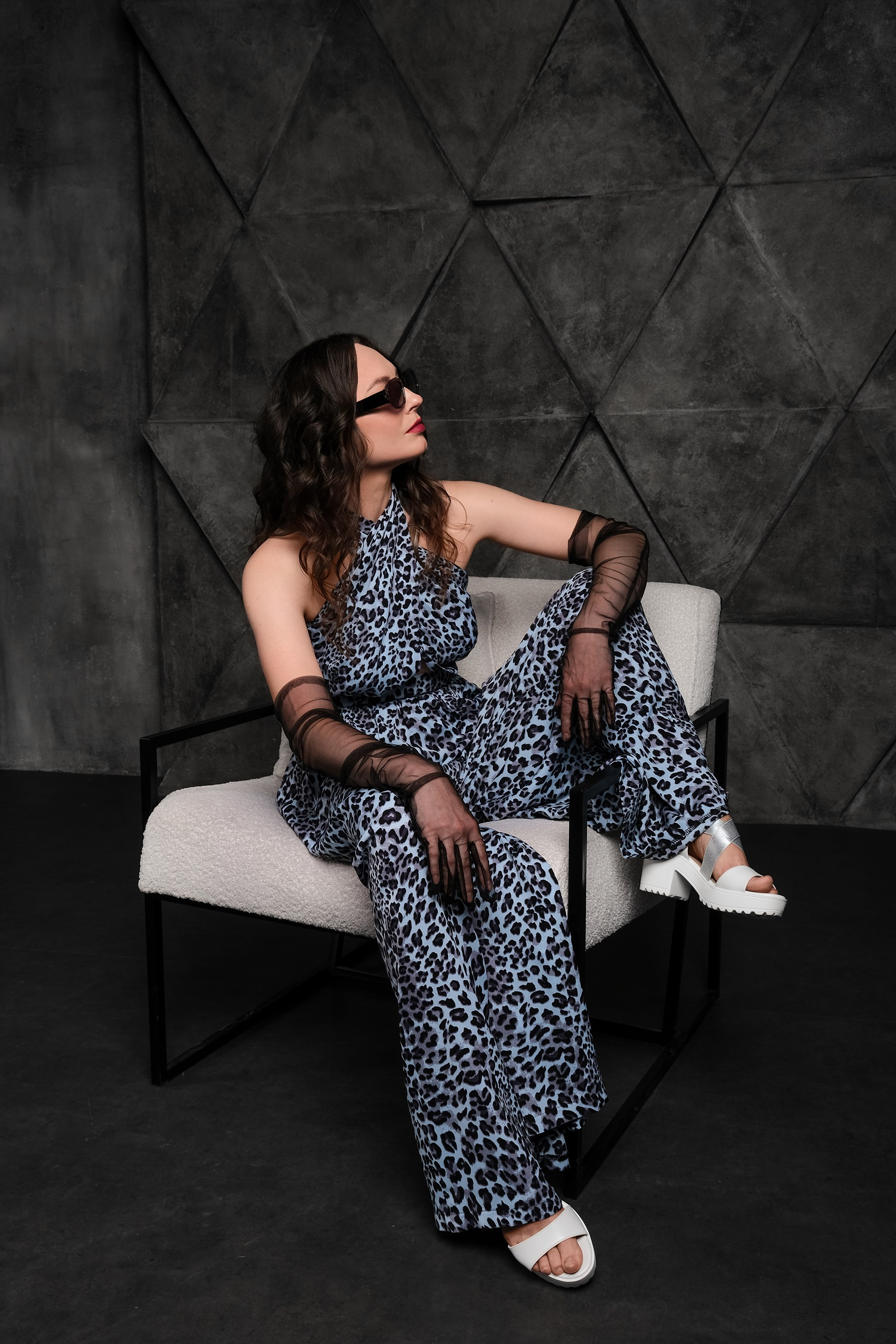 стилизованная женская фотосессия в студии. Девушка в синем комбинезоне сидит в кресле в стильных очках