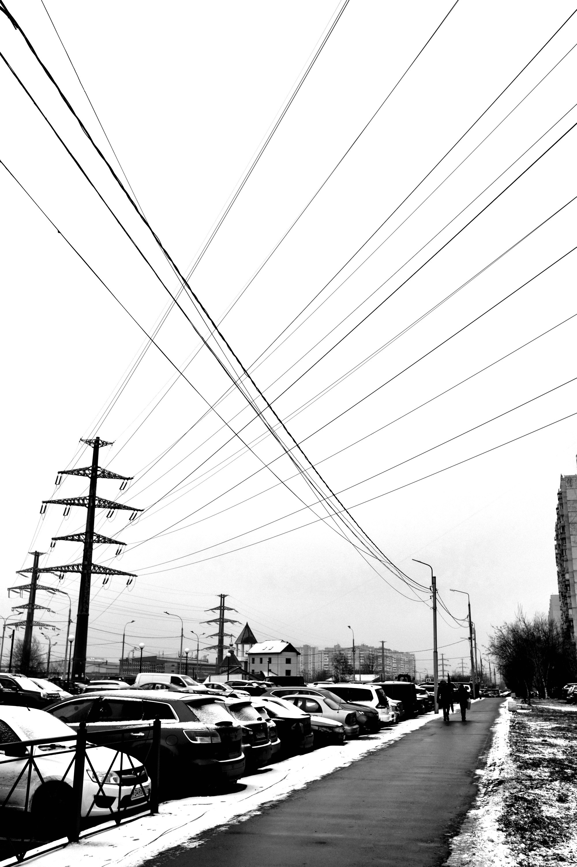 Черно-белое фото городской улицы с линиями электропередач, парковка автомобилей, силуэты людей, зимний пейзаж