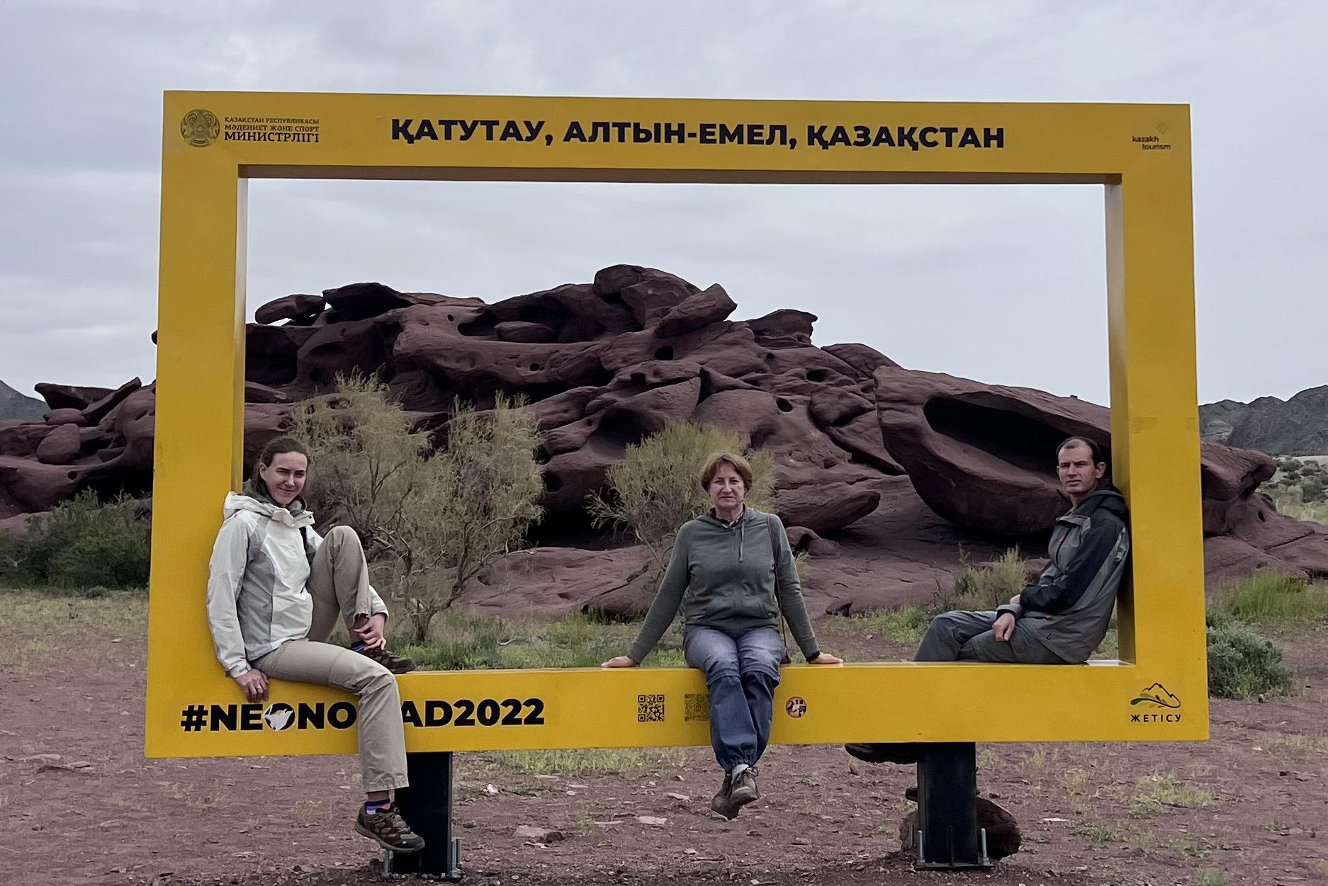 Группа из четырёх человек улыбается и позирует на фоне деревянной арки с надписью "Медвежий водопад". Люди выглядят счастливыми, одеты в светлую и спортивную одежду, очевидно, находятся в походе. За аркой видны склоны с зеленью и каменистая тропа. Атмосфера весёлая и дружелюбная, подчеркивает любовь к природе и приключениям.