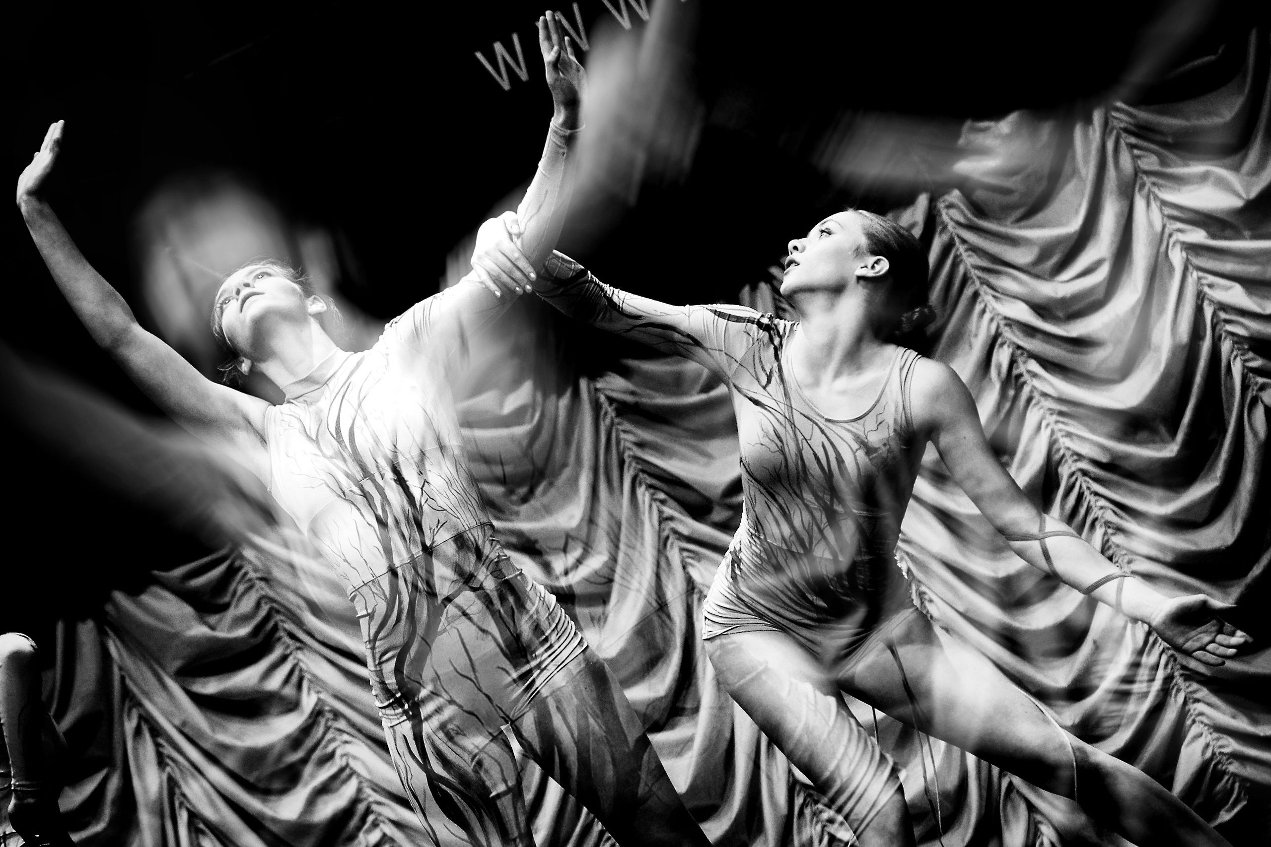 Два танцора в светлых костюмах с поднятыми руками и эффектом размытия, задник с драпировкой ткани, театральный танец