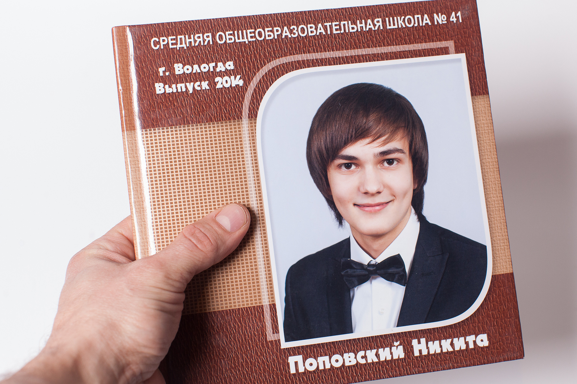 Рука держит коричневый квадратный выпускной альбом с портретом выпускника 11 класса на сером-голубом фоне.