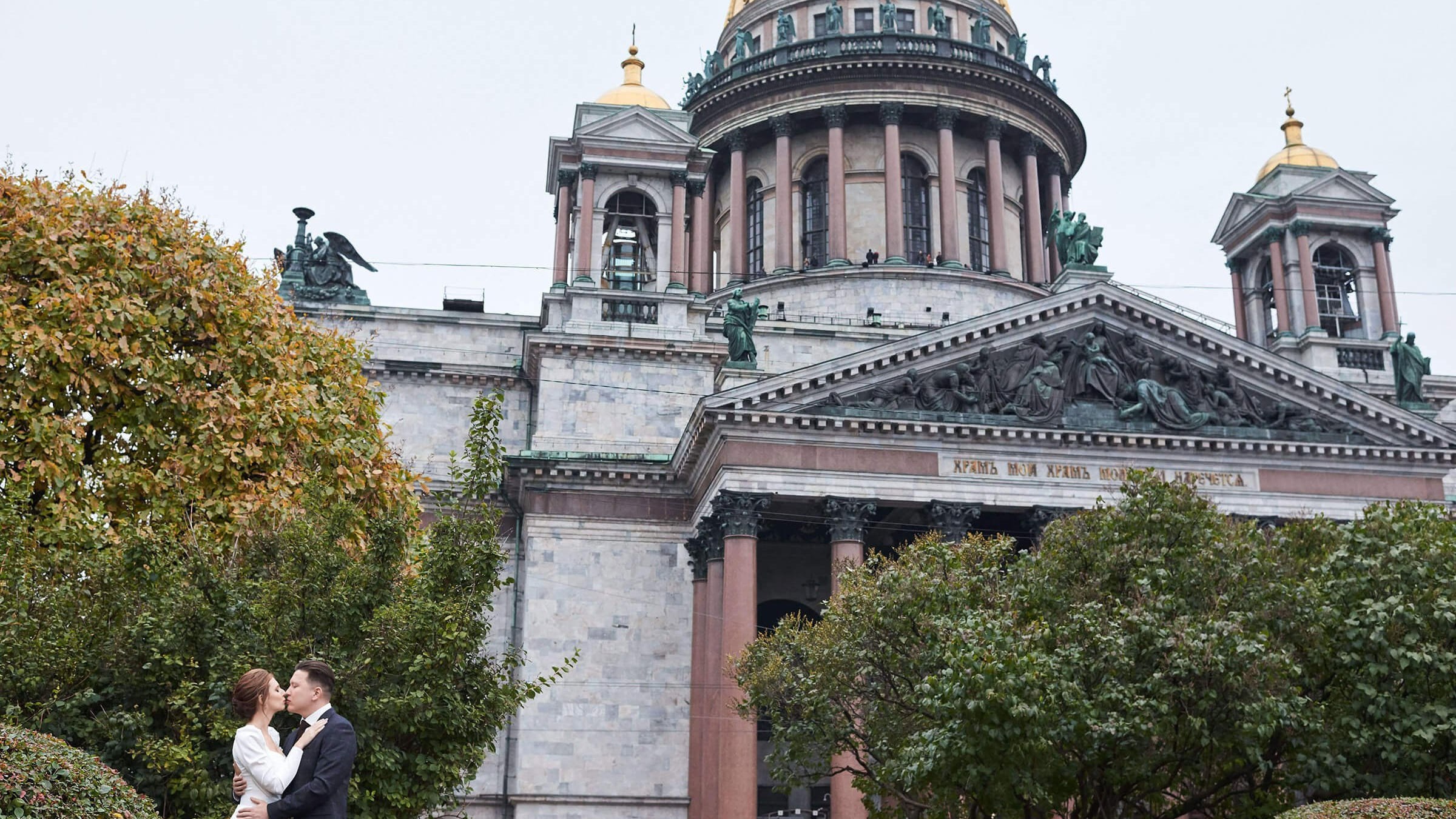 Пара обнимаетя на площади перед Исаакиевским собором. Архитектурные детали собора создают величественный фон. Дневной свет подчёркивает контрасты: белоснежные одежды невесты на фоне зеленых кустов. Композиция подчёркивает масштаб события и красоту городского пейзажа.