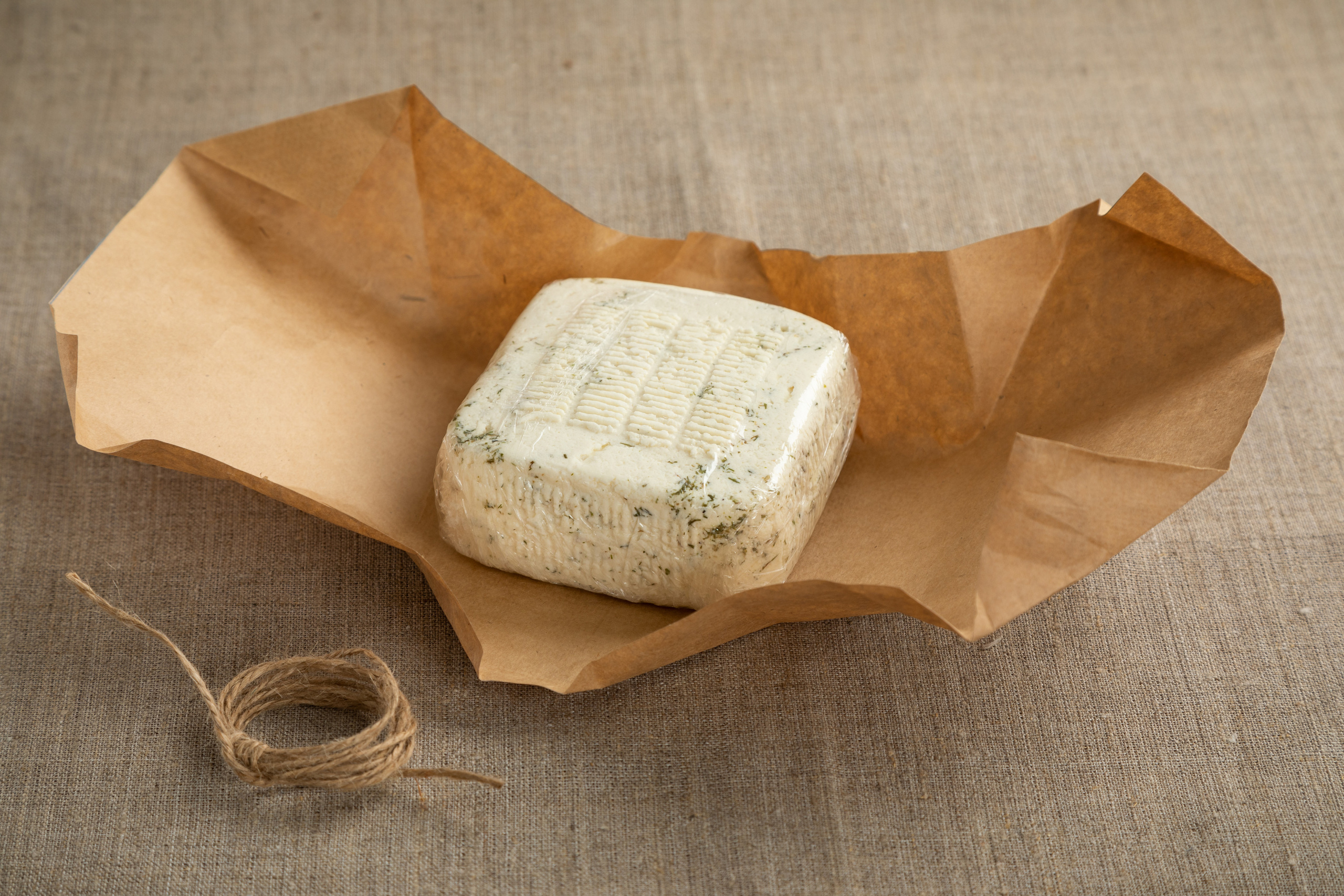 Головка адыгейского сыра с сушеным укропом развернутой упаковке из крафтовой бумаги.