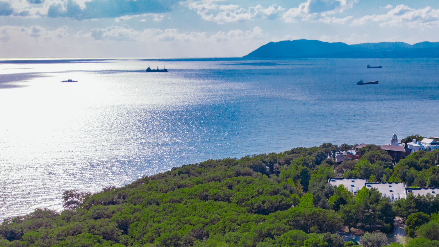 Солнце освещает пейзаж, голубое море на заднем плане, футбольное поле и здания детского лагеря на первом плане, вид с воздуха