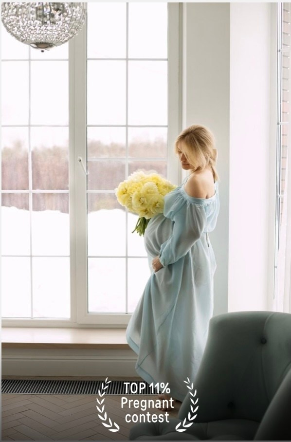 Беременная женщина в голубом платье с букетом желтых пионов стоит у большого окна.
