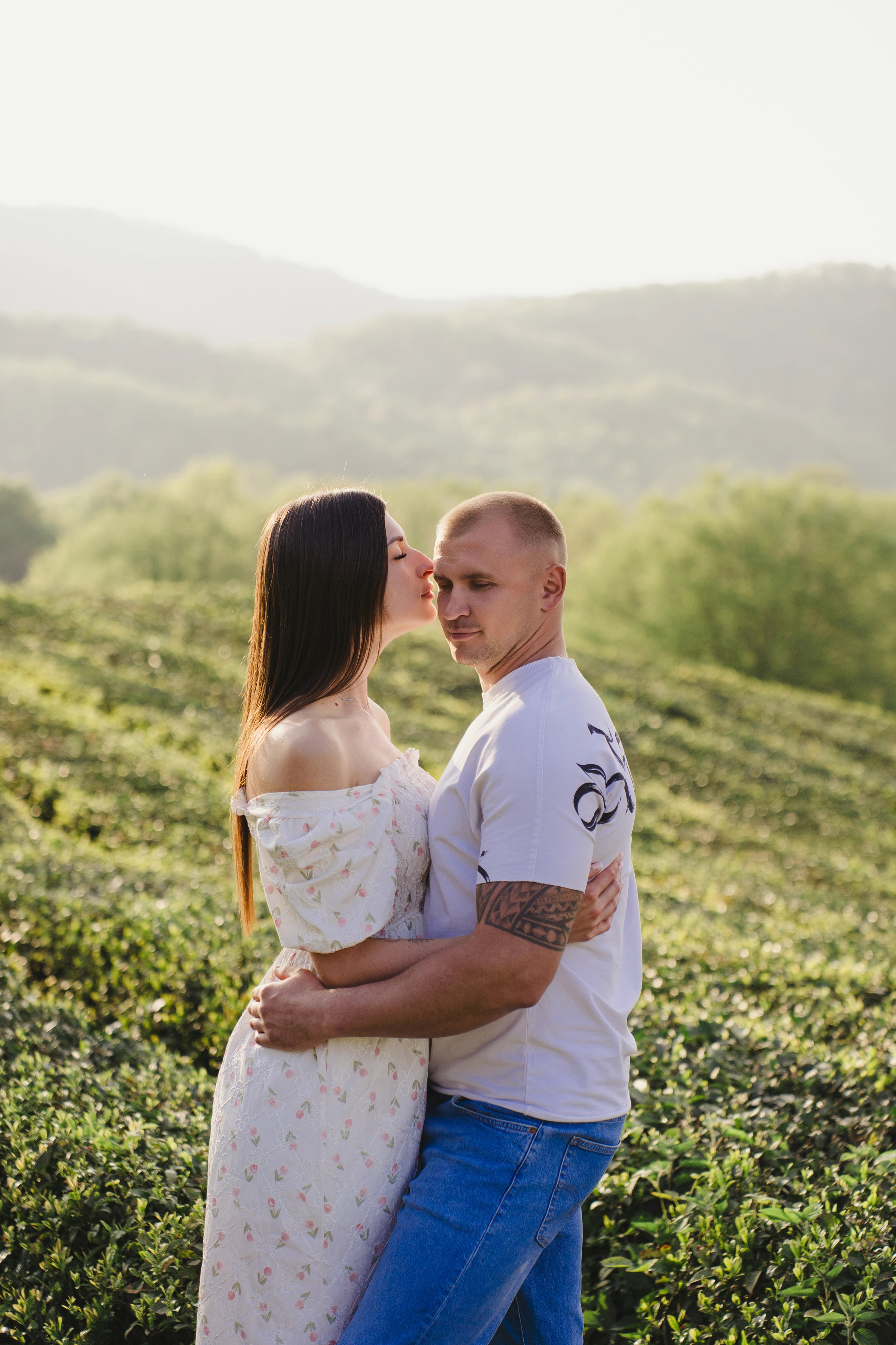 Это изображение мужчины и женщины, предположительно пары, стоящих в поле с высокой травой. Женщина стоит слева от мужчины. Она обнимает его за талию и наклоняется, чтобы поцеловать в лоб. Мужчина тоже обнимает ее за талию. Он смотрит на нее сверху вниз с улыбкой на лице. Фон изображения представляет собой размытые зеленые деревья и горы вдалеке.