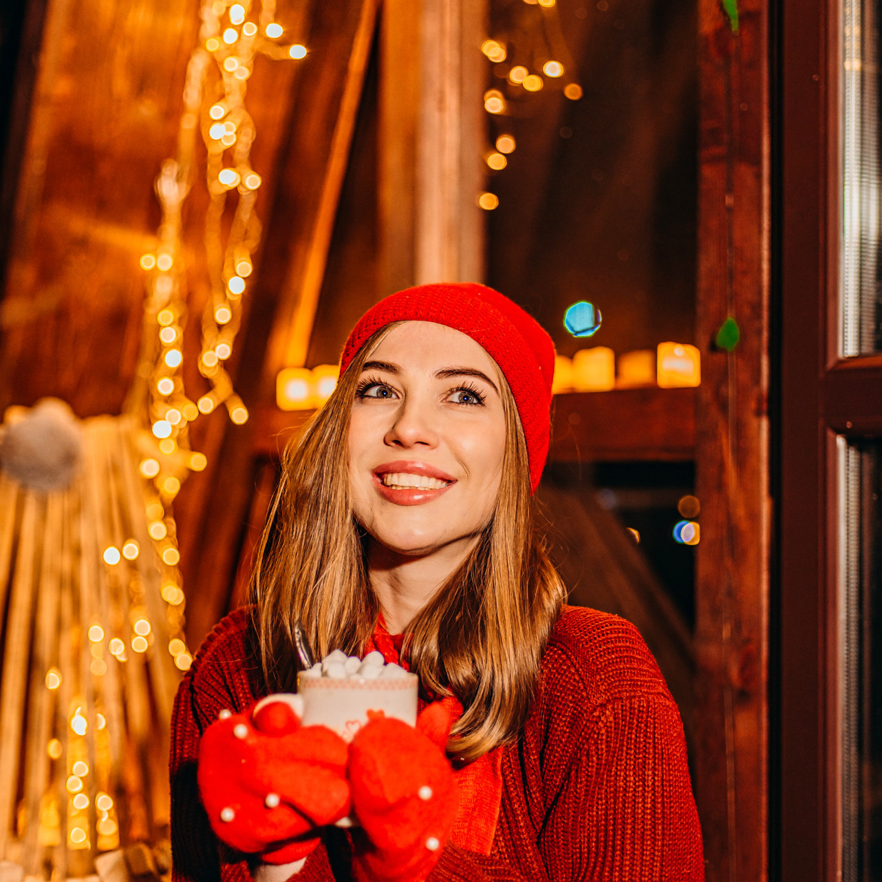 Новогодний фотопроект в Иркутске Девушка в красной шапке и варежках на фоне гирлянд держит в руках кружку с горячим шоколадом и маршмелоу