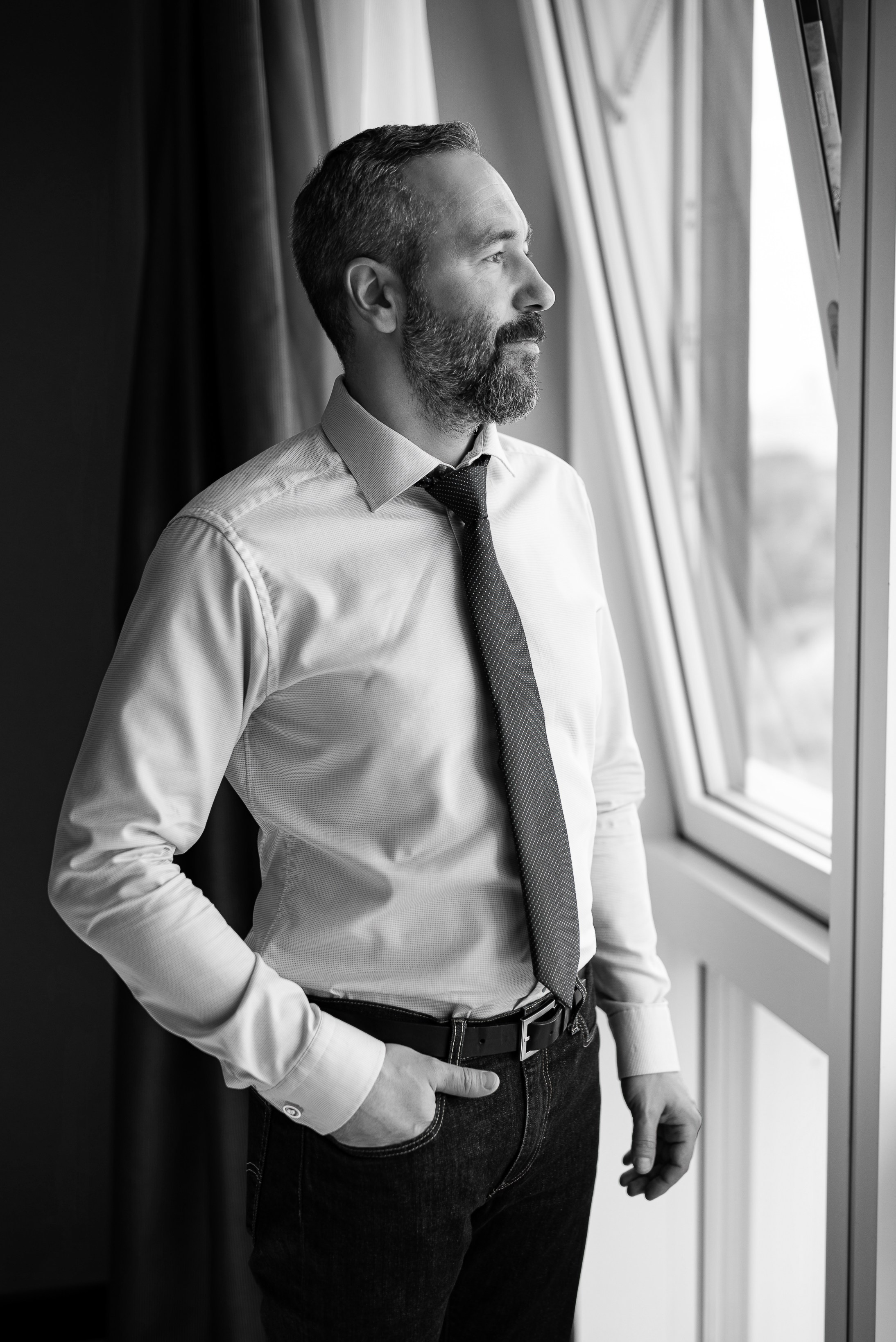 Жених на свадебной фотосессии в отеле. Жених стоит возле окна и ждёт невесту. Черно-белая фотография, свадебная фотосессия, черно-белая фотография жениха.