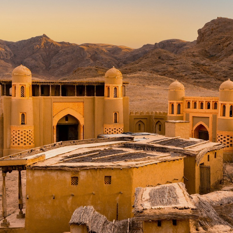 Ярко-бирюзовое озеро Иссык недалеко от Алматы окружено густыми хвойными лесами на фоне высоких гор. На переднем плане видны деревья и травянистая растительность, подчёркивающие природную красоту пейзажа. Чистое голубое небо добавляет яркости сцене.