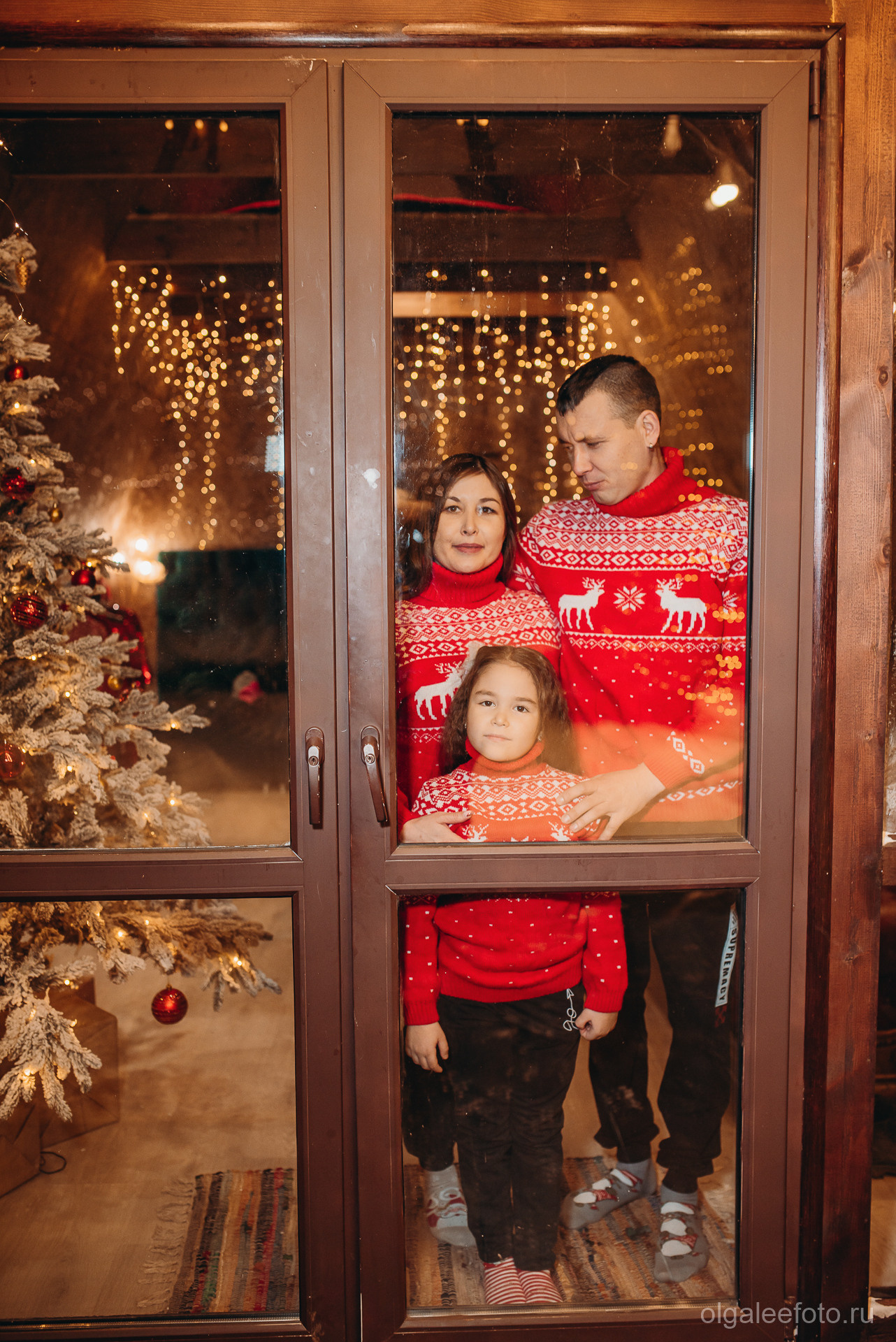 Семейная фотосессия в Иркутске Мама папа и дочь семья в красных новогодних свитерах за окном новогодней студии