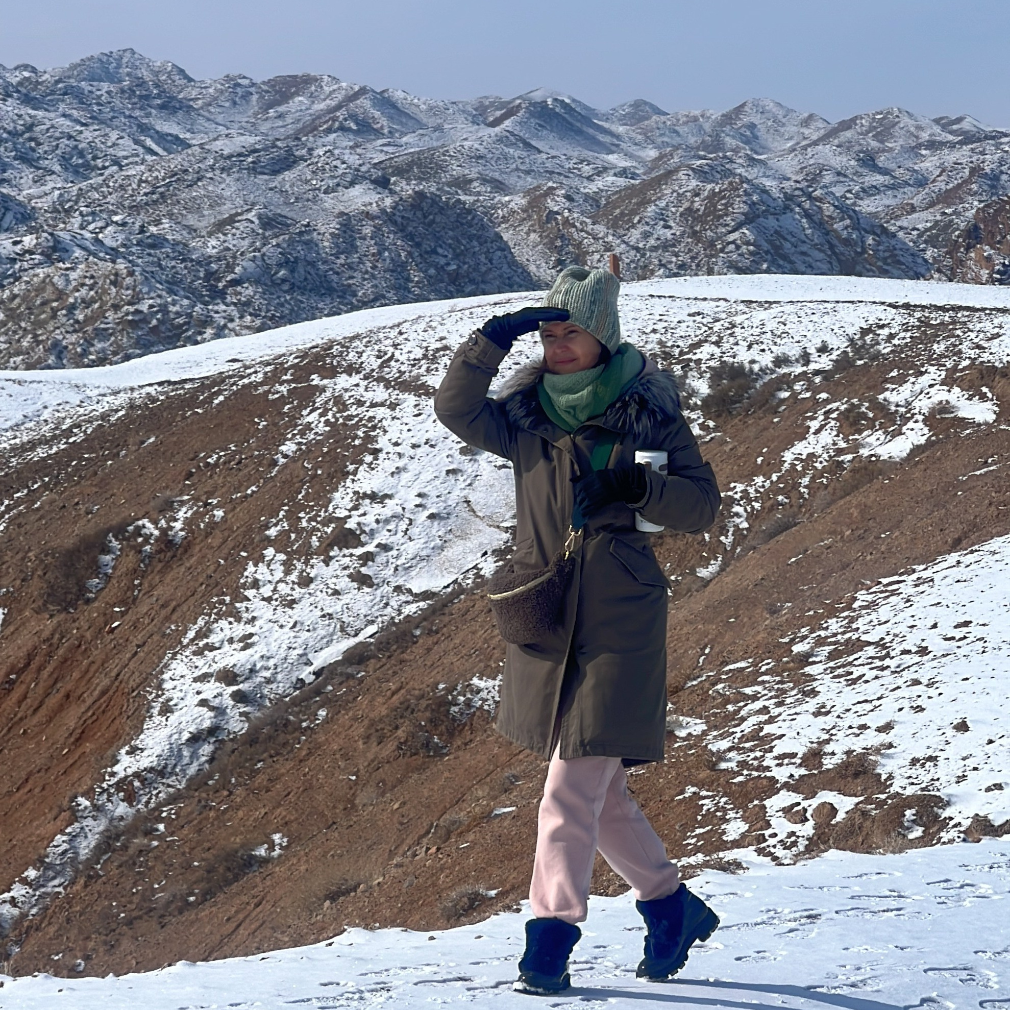 Женщина, одетая в теплую зимнюю одежду, стоит на заснеженном холме и прикрывает глаза рукой от яркого солнца, словно вглядываясь вдаль. На заднем плане видны холмы и горы, покрытые снегом, под ярко-голубым небом. Ландшафт Лунного каньона в Алматы в Казахстане вокруг сочетает белый снег и оголенные участки коричневой земли.