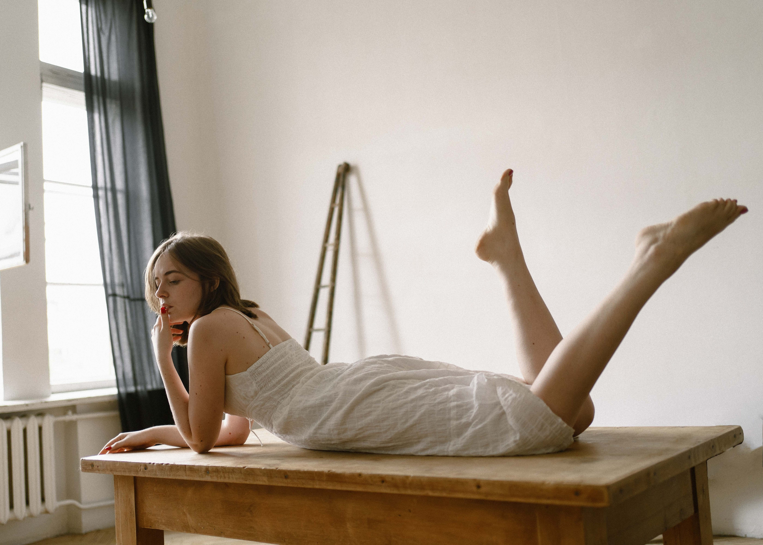 Фотография, где девушка лежит на столе, машет ногами. Атмосфера на фото спокойная, меланхоличная. Девушка одета в белый сарафан, у нее короткая стрижка, задумчивый взгляд.