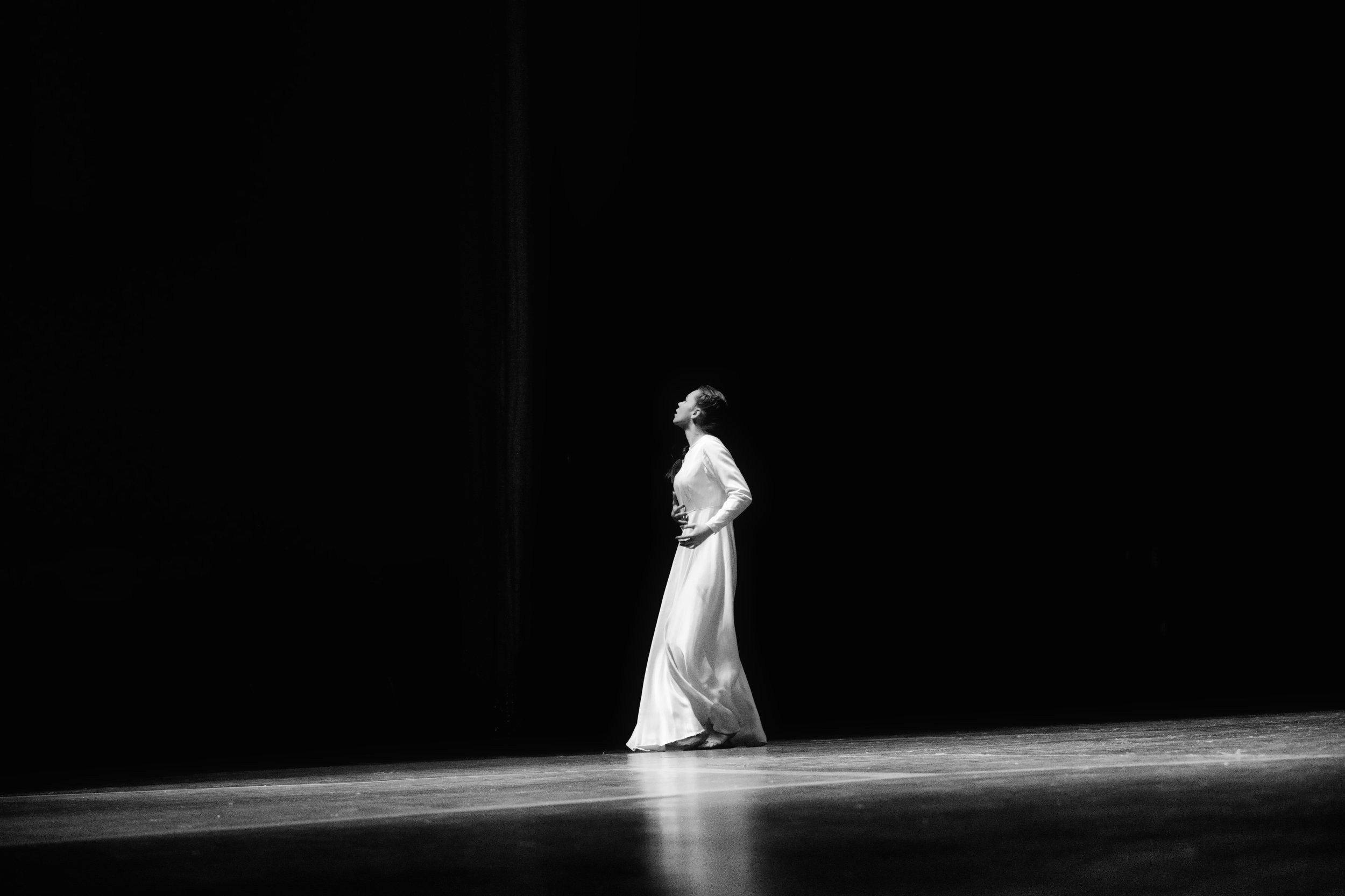 Черно-белая театральная фотография: балерина в длинном белом платье стоит на сцене в профиль, голова поднята вверх, драматичное освещение, темный фон, минималистичная композиция