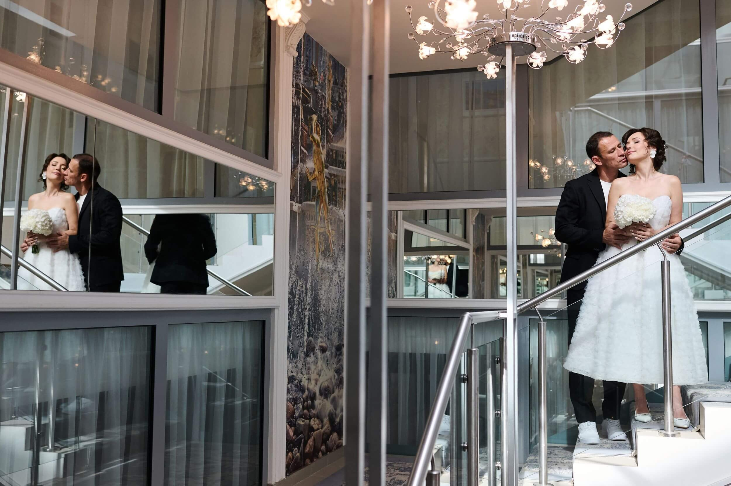 Молодожёны на парадной лестнице Отдела‑дворца РАГС Петродворцового района в Петергофе. Отражение в зеркалах, светлое оформление и естественный свет создают элегантный фон для свадебного снимка. Классический ракурс, подчёркивающий торжественность момента.