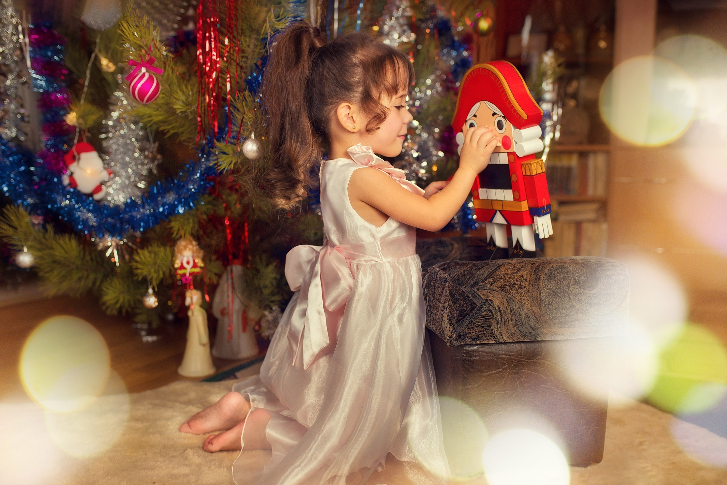 Девочка в нарядном платье возле новогодней ёлки играет с игрушкой Щелкунчик