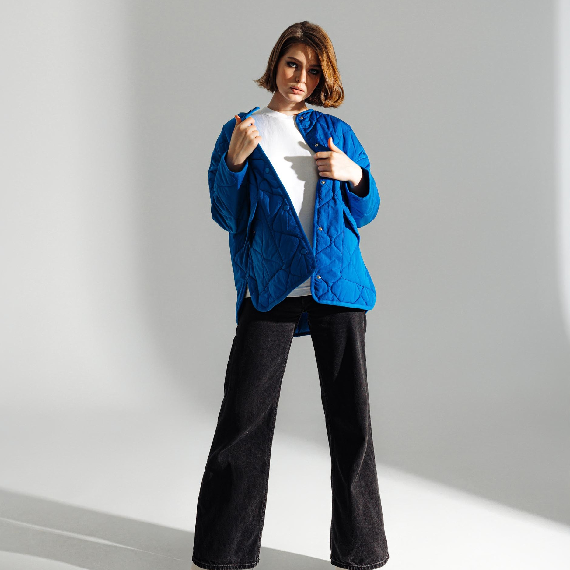 Молодая девушка в джинсах клеш и синей куртке стоит на фоне циклорамы