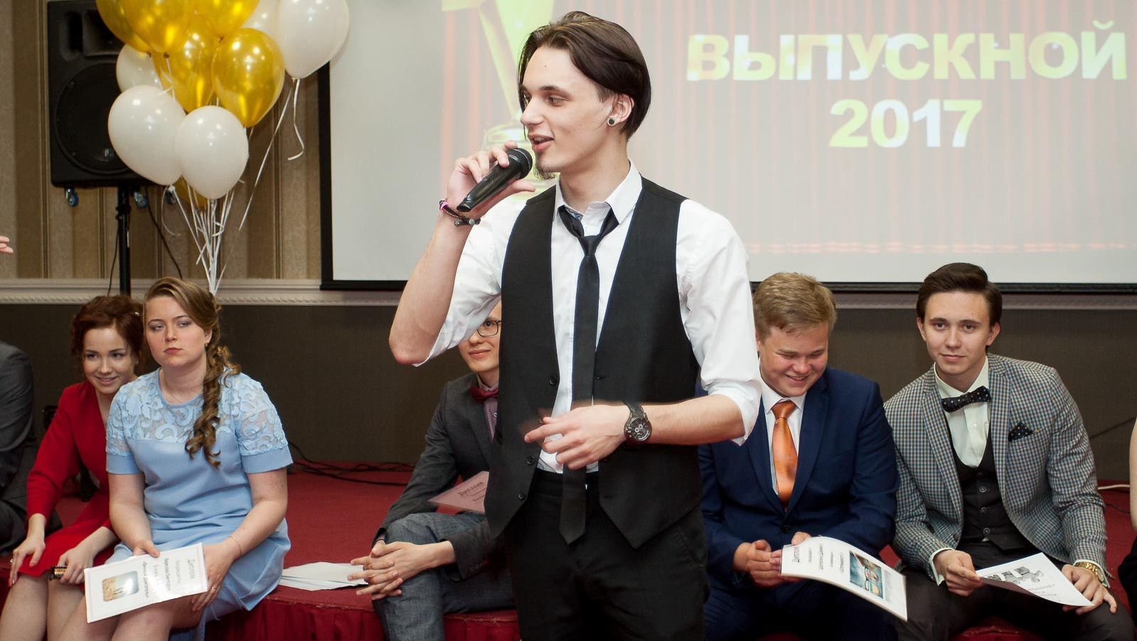 Выпускник в тройке стоя говорит в микрофон на фоне сидящих одноклассников на сцене.