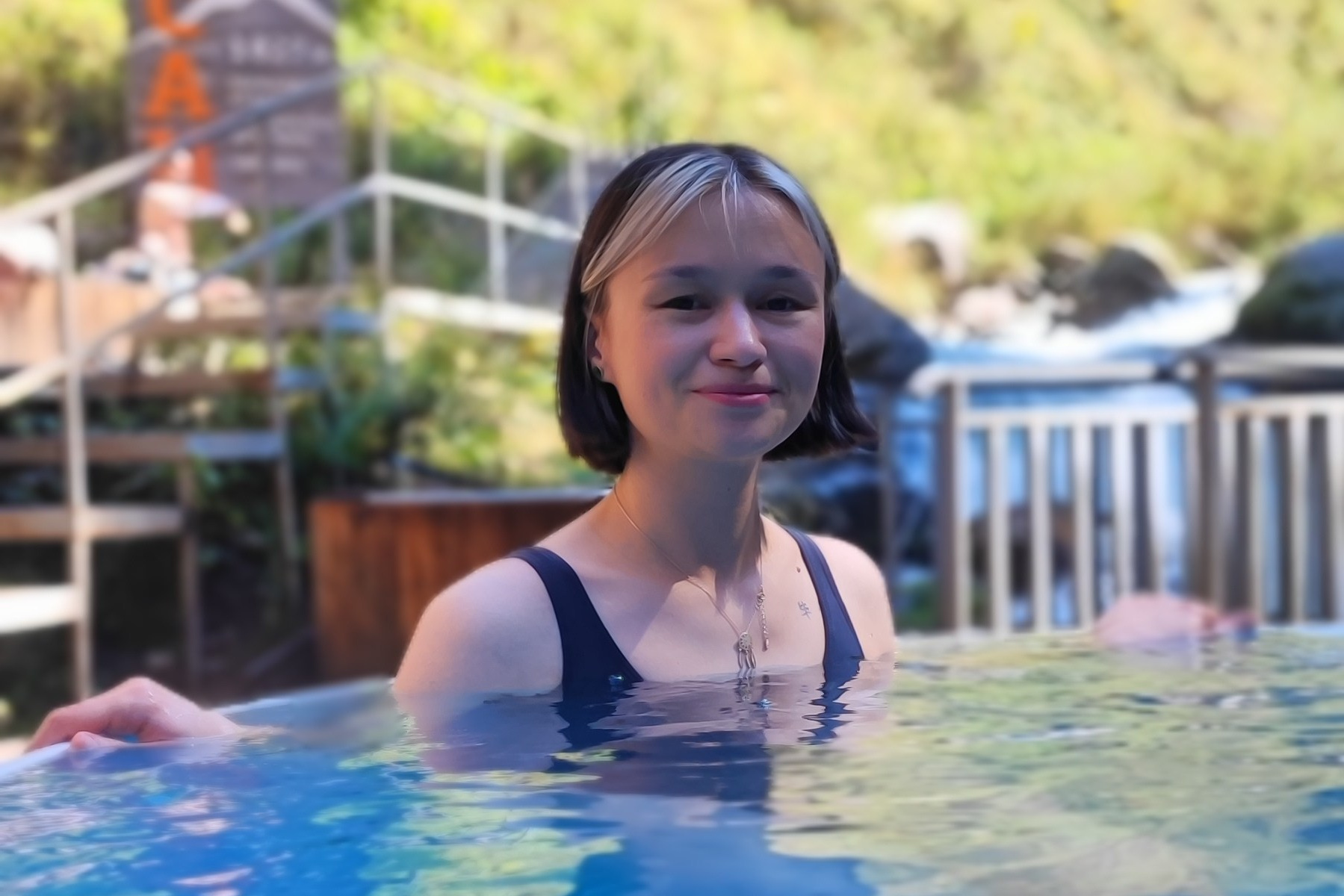 Человек позирует на фоне бирюзового озера Иссык в районе Алматы и гор. Он стоит на большом камне, одет в ярко-зеленую толстовку, черные штаны и светлую обувь, а также оранжевую панаму. В руках держит очки. За ним виднеются деревья, лесистые склоны и горные вершины под облачным небом. Атмосфера спокойная и природная.