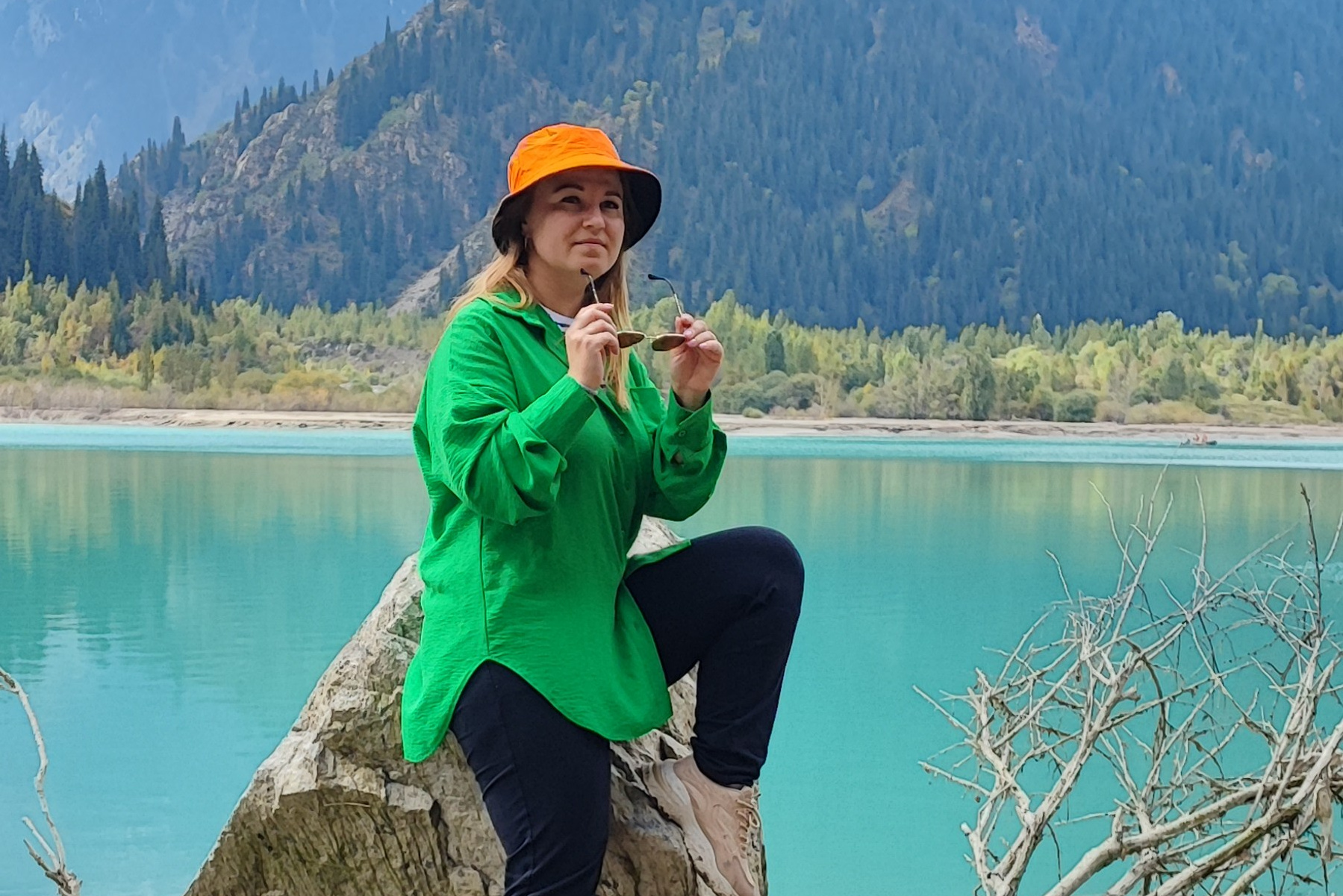 Человек позирует на фоне бирюзового озера Иссык в районе Алматы и гор. Он стоит на большом камне, одет в ярко-зеленую толстовку, черные штаны и светлую обувь, а также оранжевую панаму. В руках держит очки. За ним виднеются деревья, лесистые склоны и горные вершины под облачным небом. Атмосфера спокойная и природная.