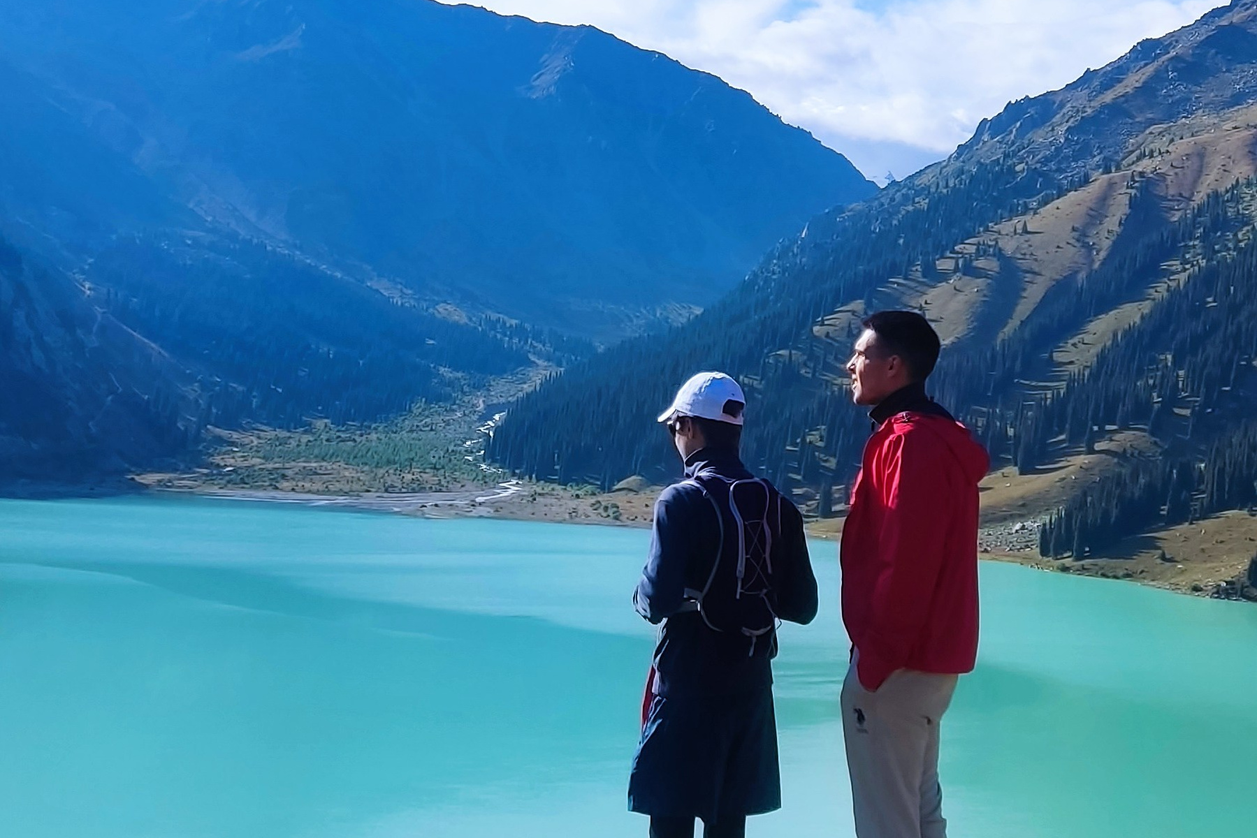 Человек позирует на фоне бирюзового озера Иссык в районе Алматы и гор. Он стоит на большом камне, одет в ярко-зеленую толстовку, черные штаны и светлую обувь, а также оранжевую панаму. В руках держит очки. За ним виднеются деревья, лесистые склоны и горные вершины под облачным небом. Атмосфера спокойная и природная.