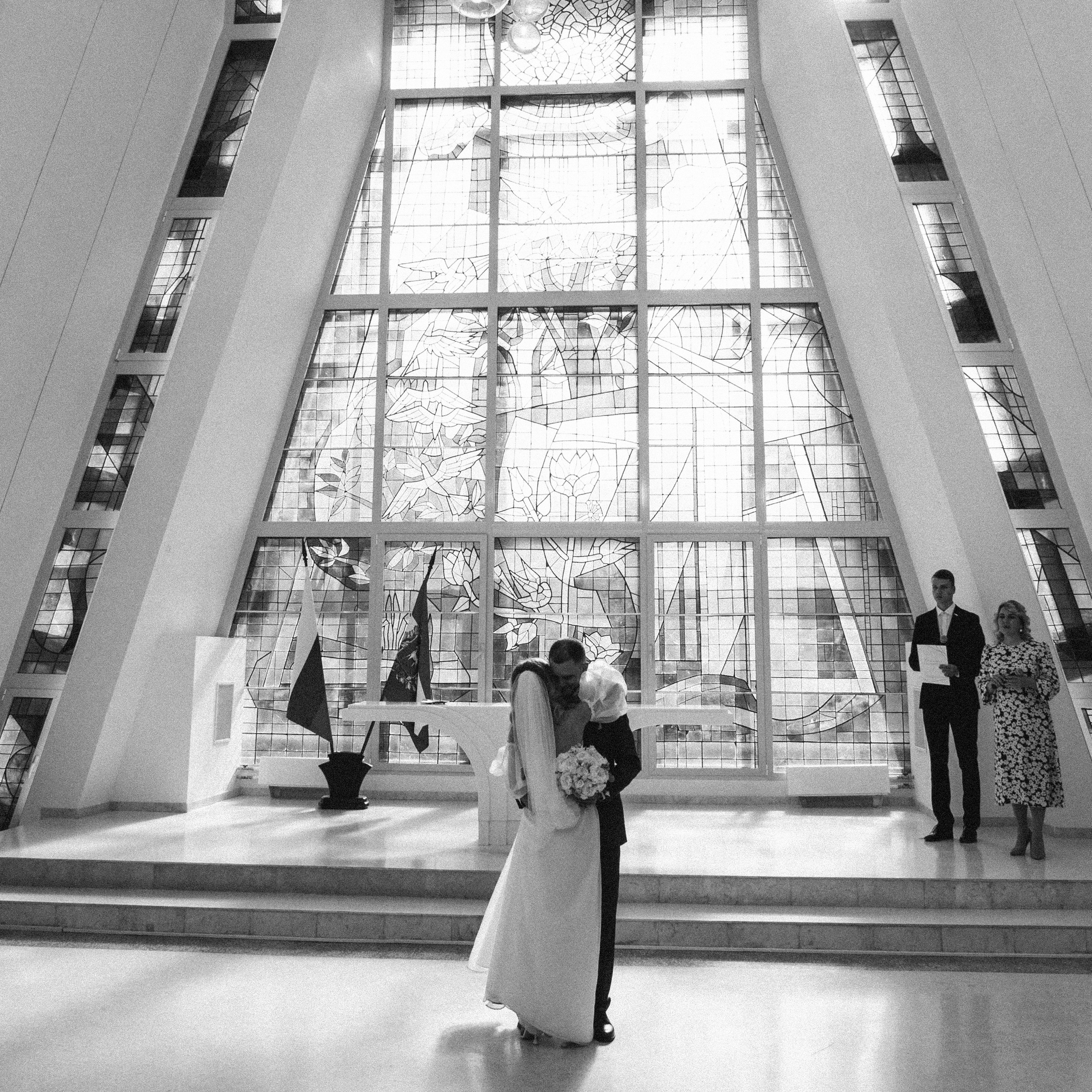 Жених с невестой нежно обнимаются стоя в зале церемоний загса на фоне советского витража