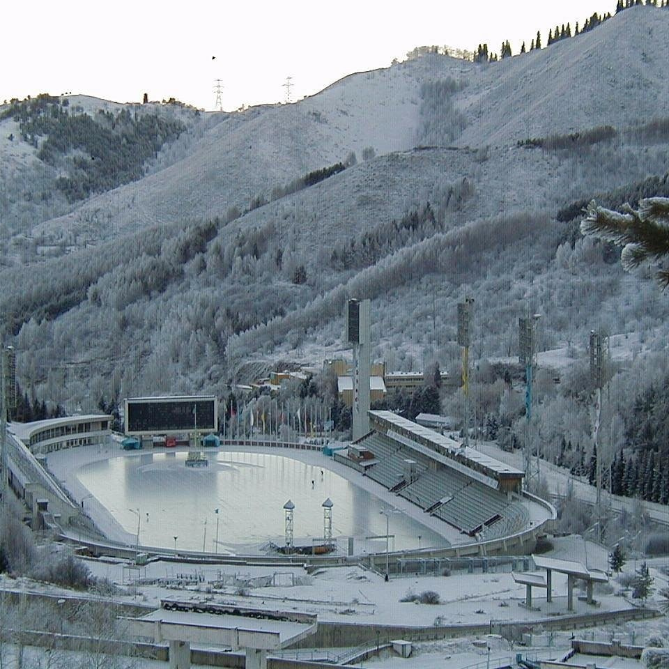 Ярко-бирюзовое озеро Иссык недалеко от Алматы окружено густыми хвойными лесами на фоне высоких гор. На переднем плане видны деревья и травянистая растительность, подчёркивающие природную красоту пейзажа. Чистое голубое небо добавляет яркости сцене.