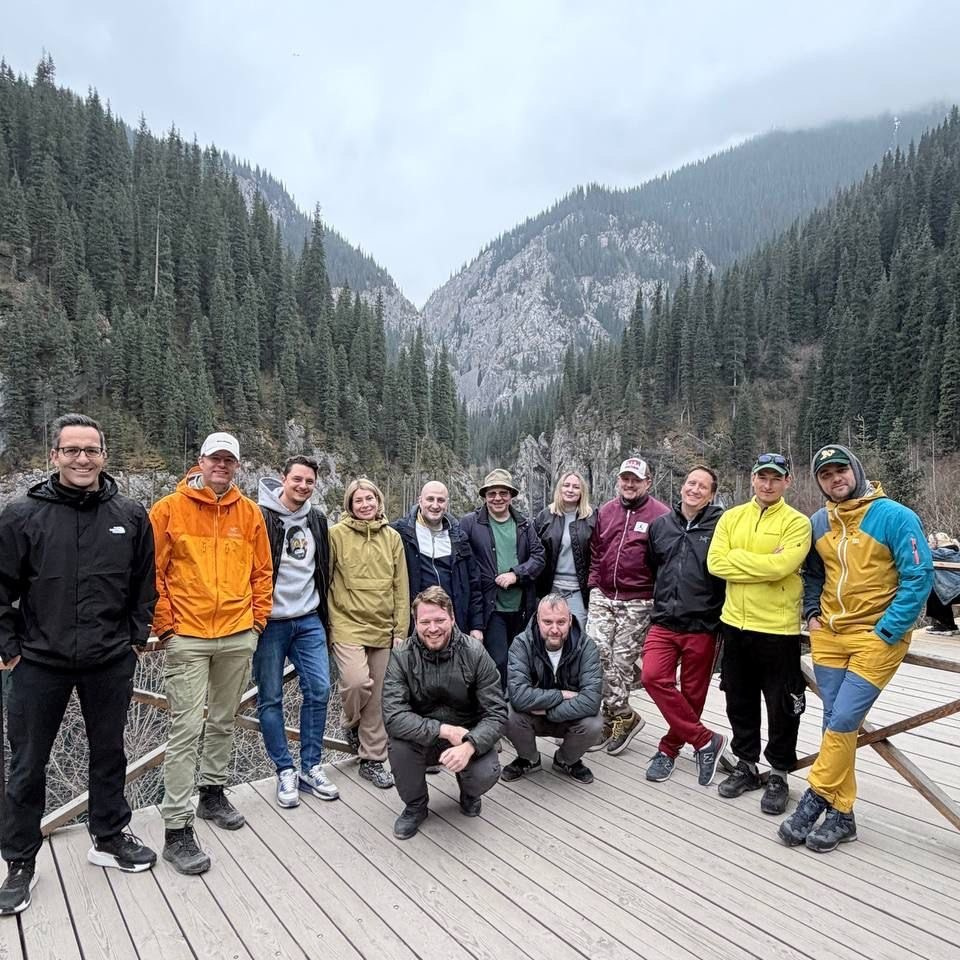 Группа людей, участников корпоративного тура Melon Travel на озеро Иссык, одетых в зимнюю одежду, позирует на фоне заснеженного пейзажа. На земле лежит снег, деревья покрыты инеем, а на заднем плане виднеется туман, создающий атмосферу спокойствия и загадочности. Участники группы улыбаются и выглядят счастливыми.