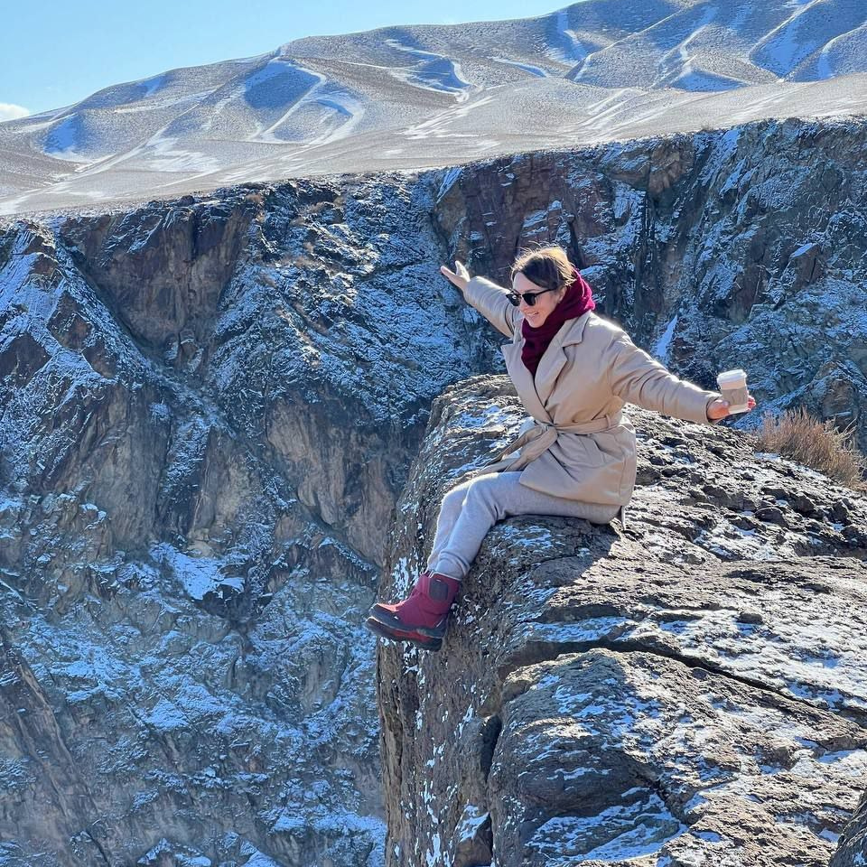 Женщина в светлом пальто, серых брюках и ярких красных ботинках сидит на краю высокой скалы Черного каньона в районе Алматы, Казахстан. На её плечах красный шарф, а в одной руке — стакан, в другой — вытянутые в стороны руки, словно она наслаждается моментом. Вокруг видно заснеженные скалы и холмы, создающие атмосферу зимнего пейзажа. На заднем плане холмы покрыты снежным покровом под ярким голубым небом.
