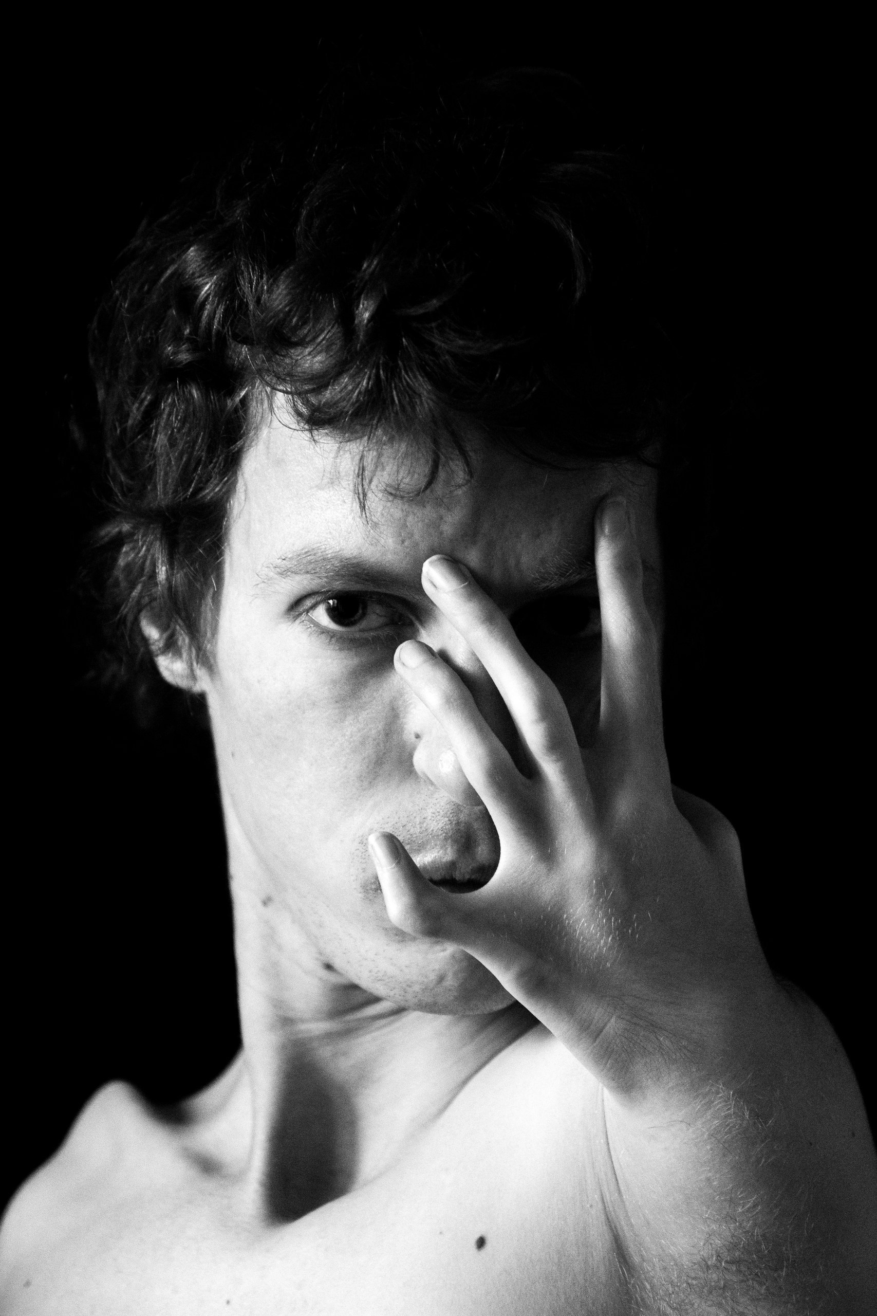 Black and white close-up art portrait of a man on a black background, gaze through his fingers, social photography by Anna Fors