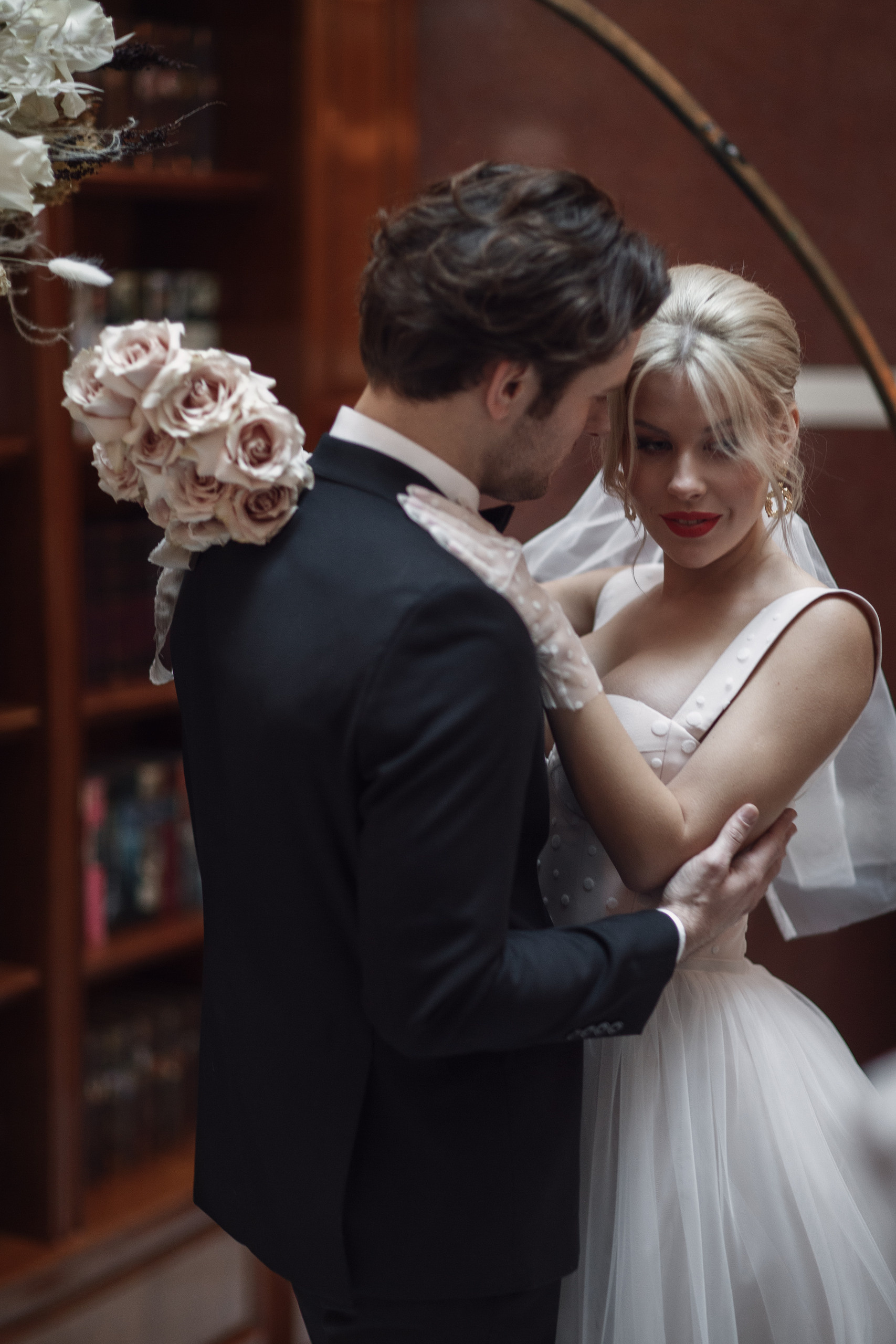 Невеста и жених у праздничной арки обнимают друг друга. У невесты в руках белые цветы, розы. 